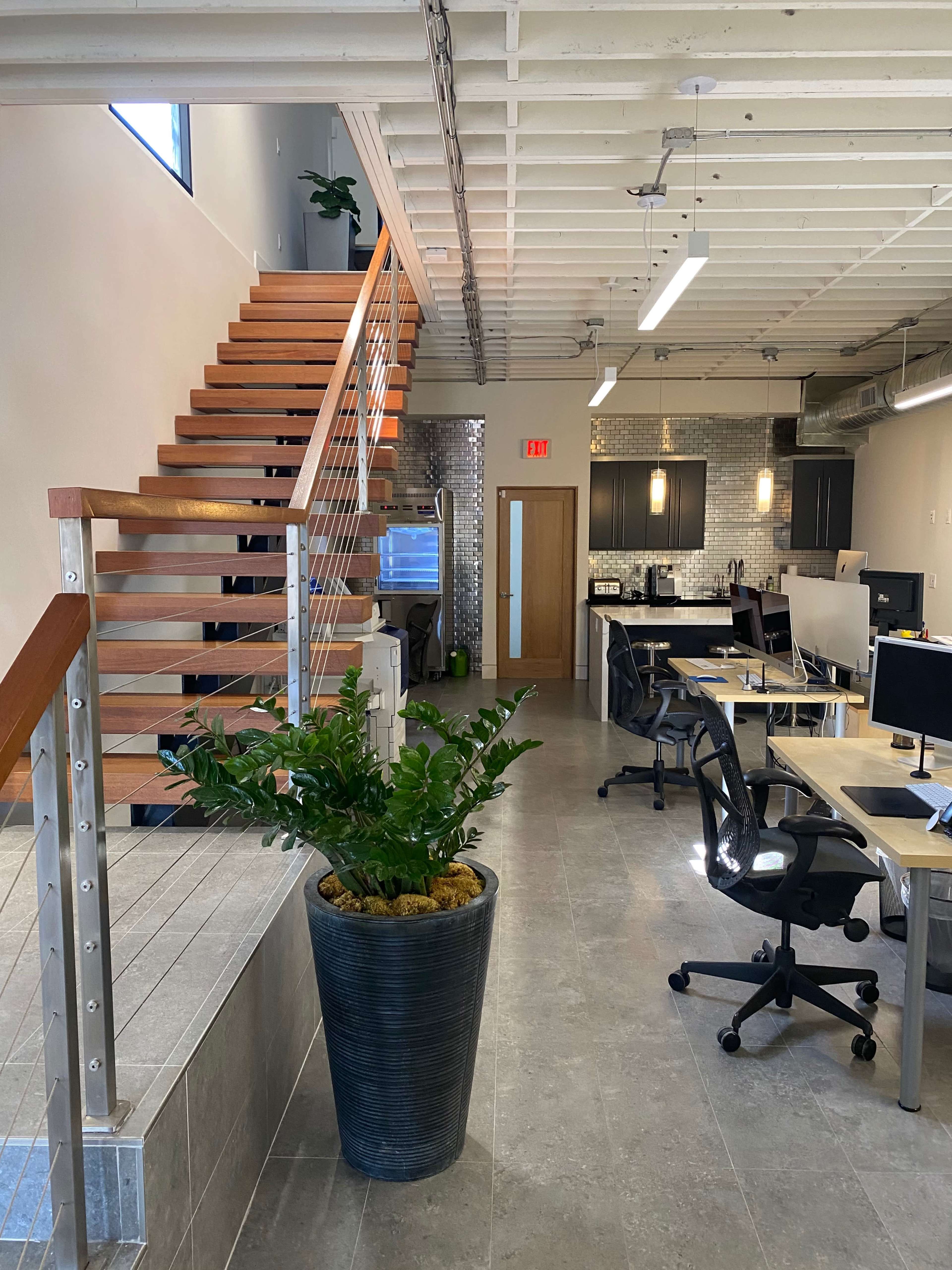 A modern office space with a staircase leading to an upper level, surrounded by desks and a large planter.