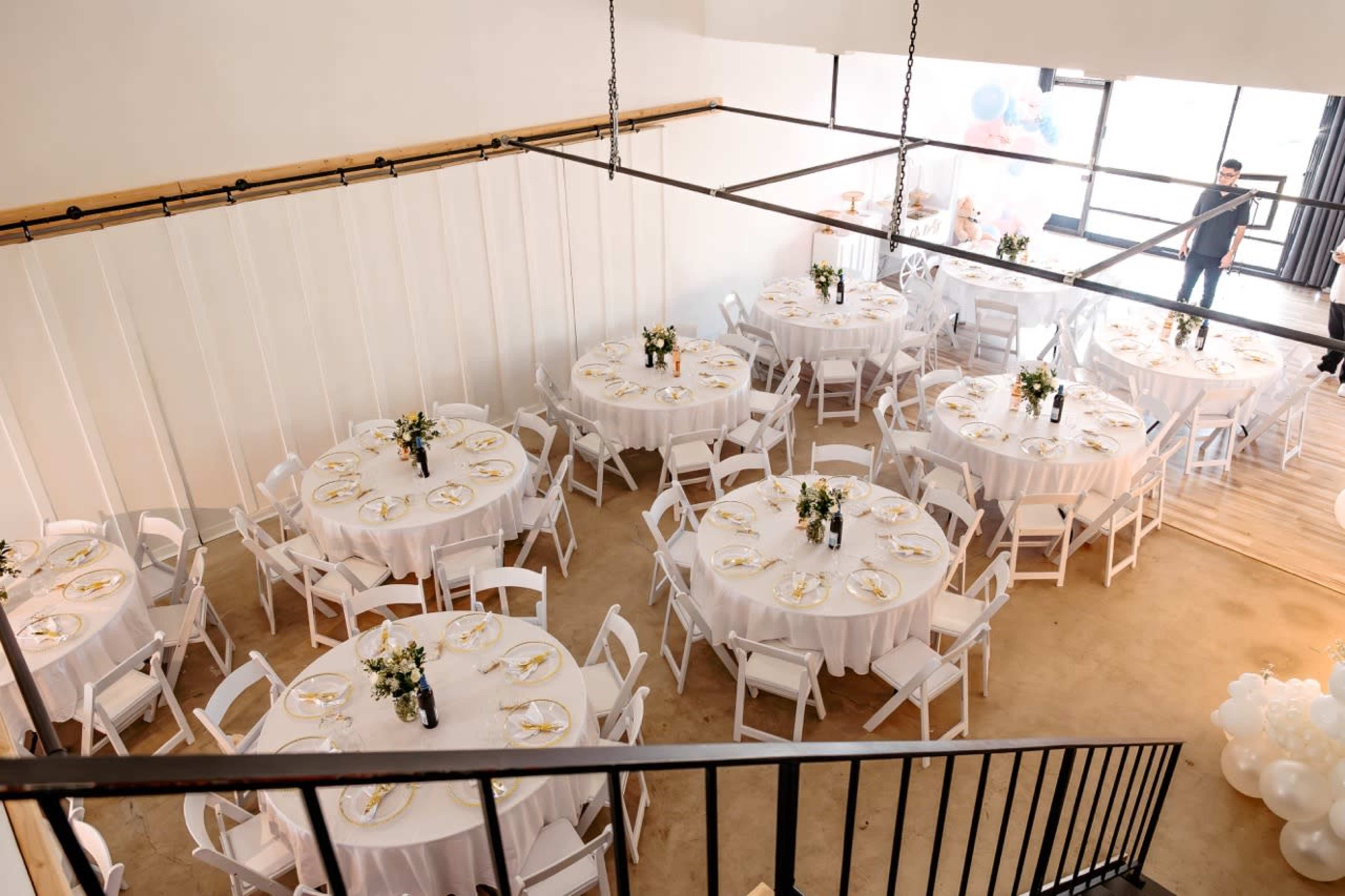 A spacious event venue with several round tables set for a formal gathering, each adorned with white tablecloths and centerpieces.