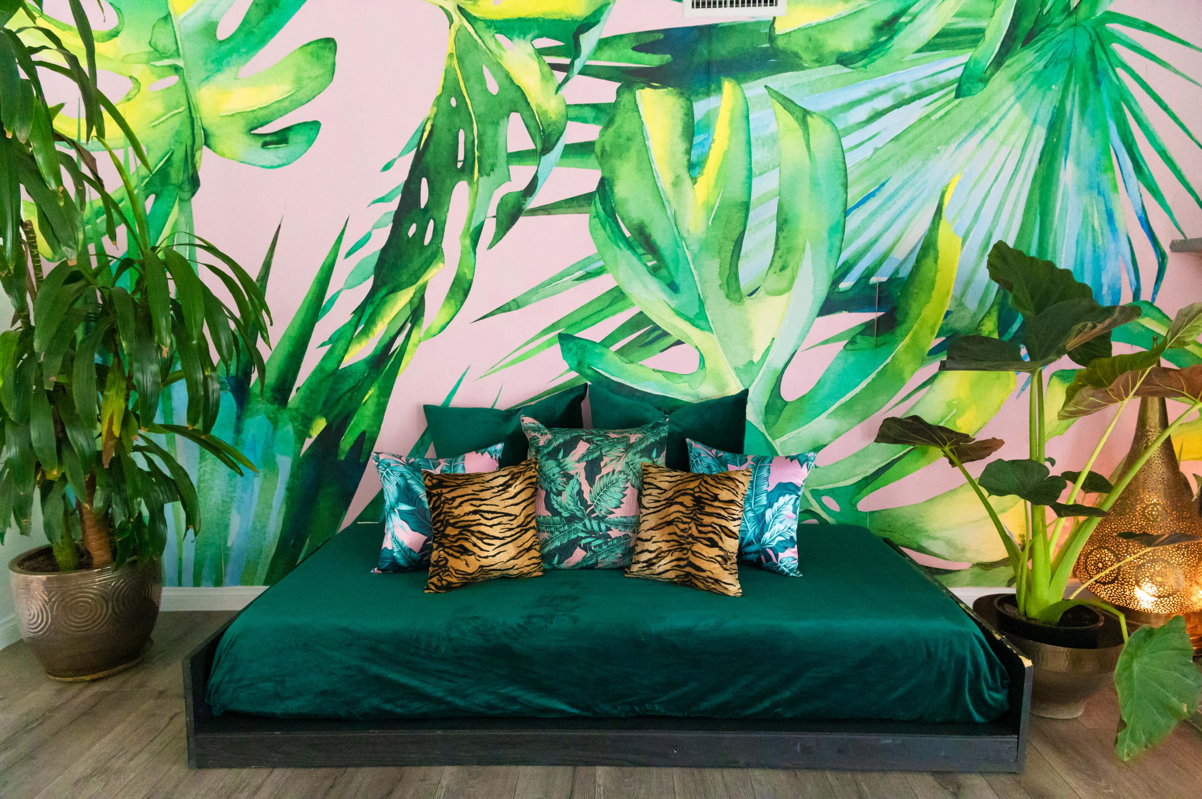 A large bed with a green velvet spread and patterned pillows is positioned in front of a vibrant tropical foliage mural on a pink wall.