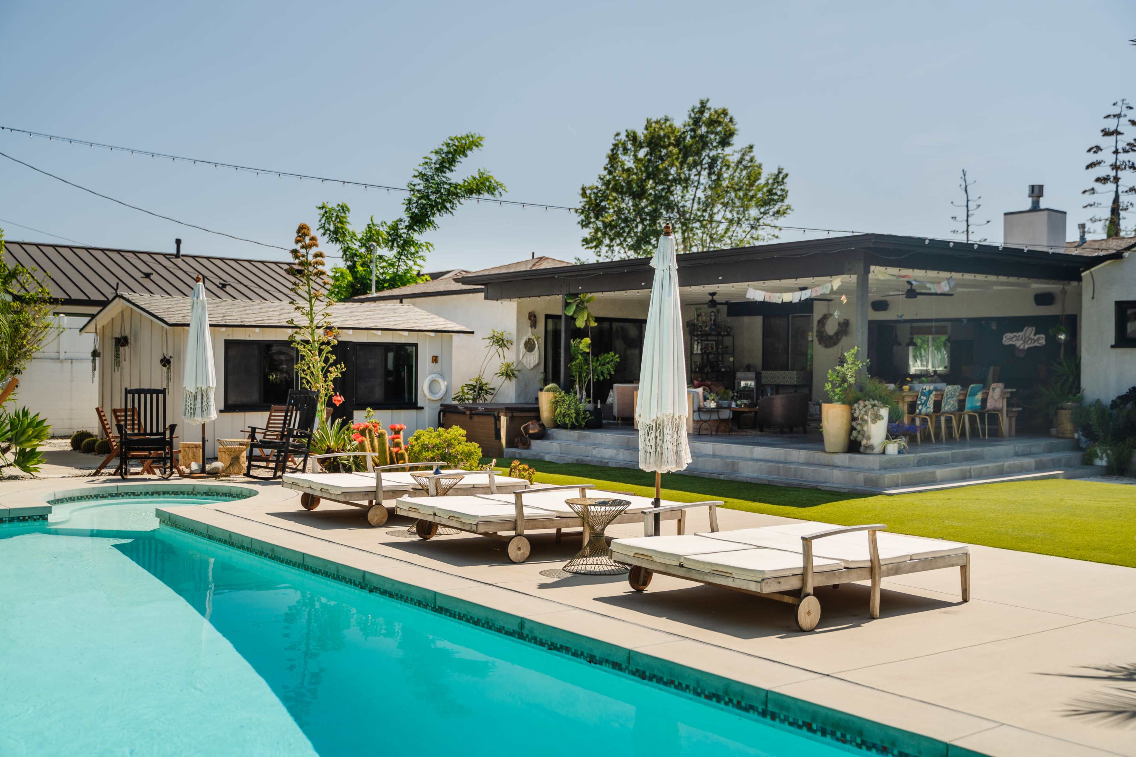 A backyard features a swimming pool with lounge chairs and a patio area displaying a covered seating space and landscaped greenery.