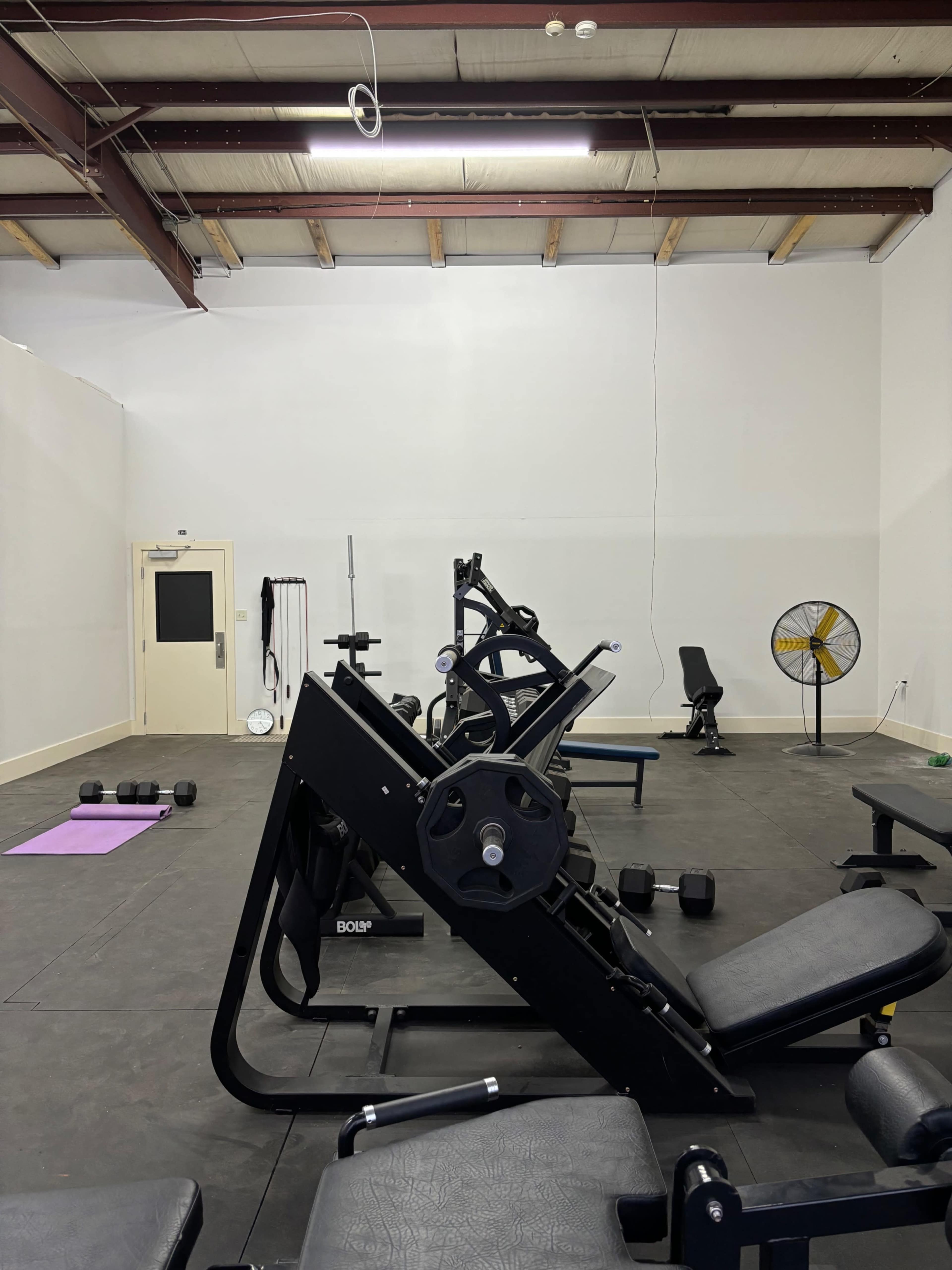 A spacious gym interior with various exercise equipment, including weights, a bench press, and a fan, against plain walls and a concrete floor.