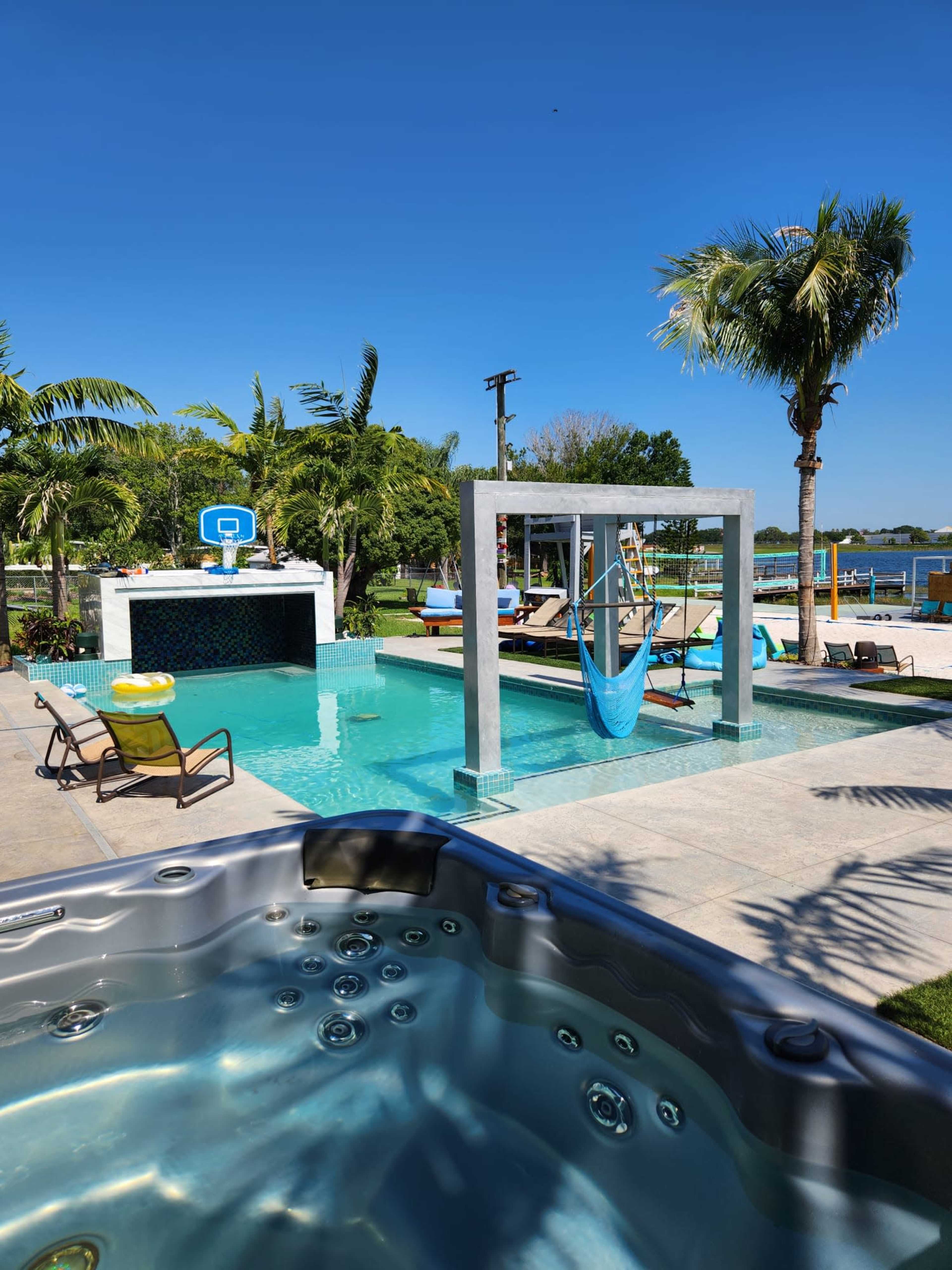 A modern backyard featuring a swimming pool, a hot tub, a hammock, and palm trees under a clear blue sky.