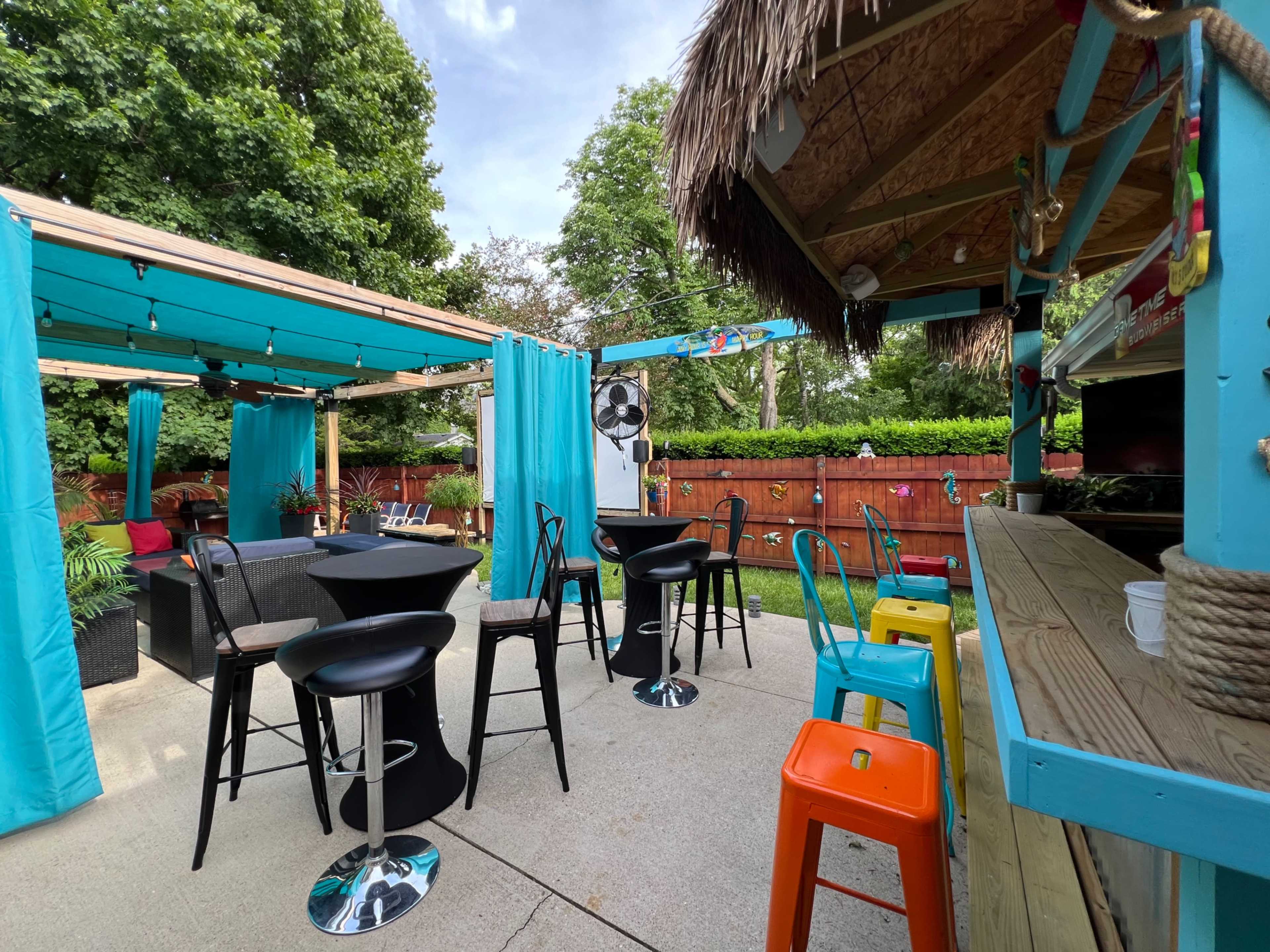 The image shows an outdoor recreational space with a bar area, colorful stools, and a seating arrangement surrounded by greenery and bright blue curtains.