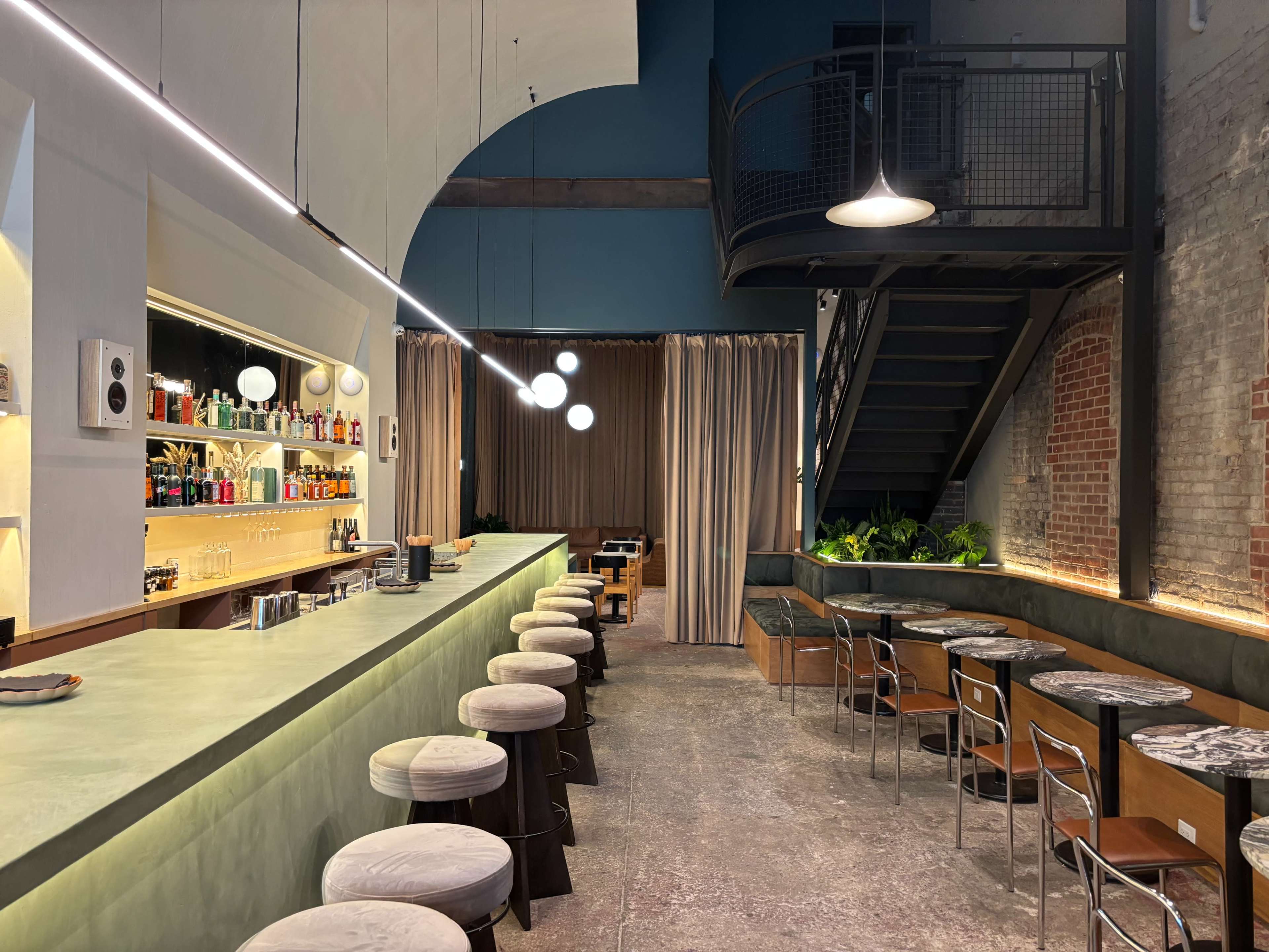 The image shows a modern bar area with a long counter, several stools, and a staircase leading to an upper level, accented by pendant lights and muted decor.