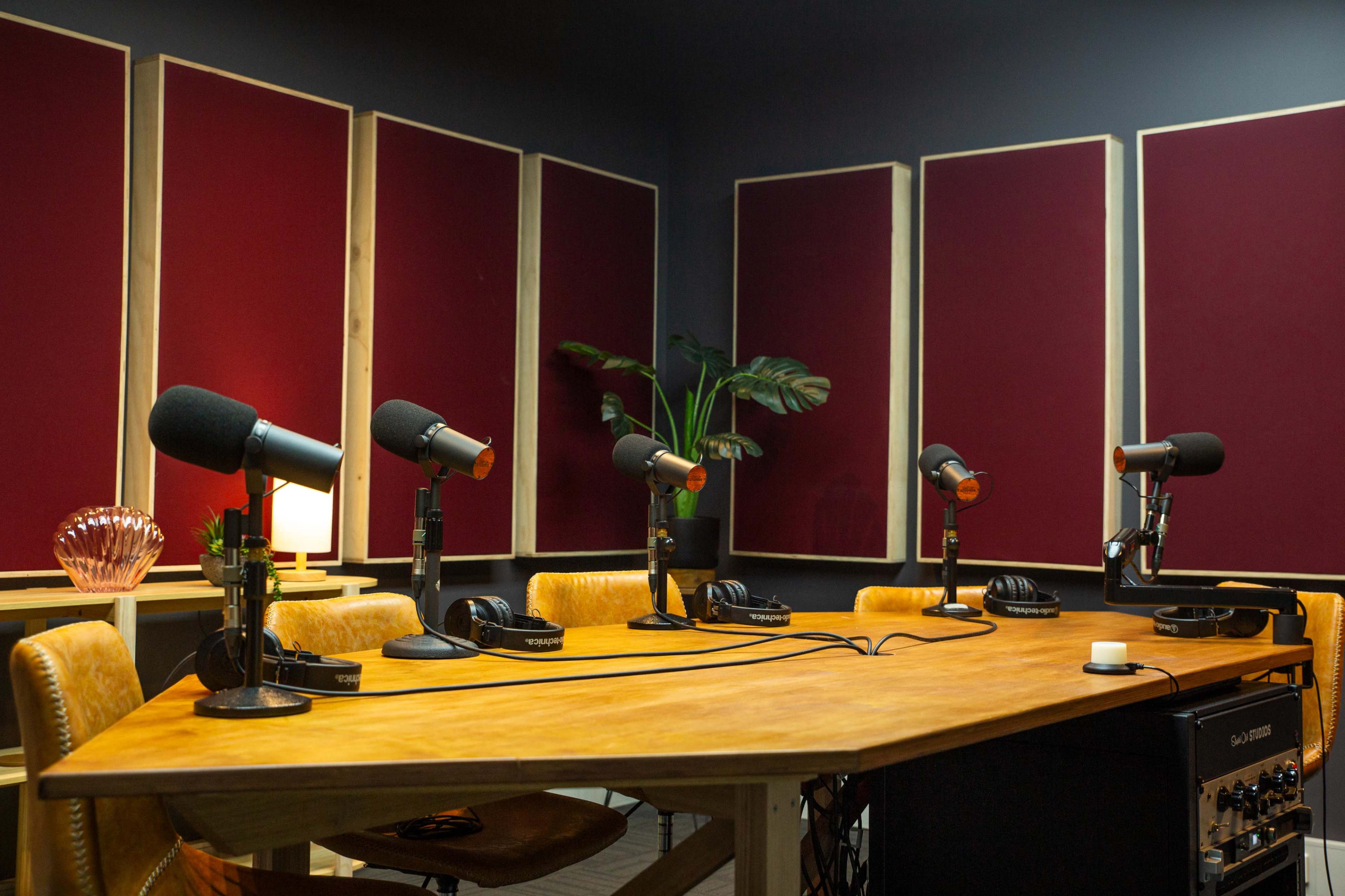 A podcast recording studio with four microphones positioned around a wooden table, surrounded by soundproof panels and minimal furnishings.