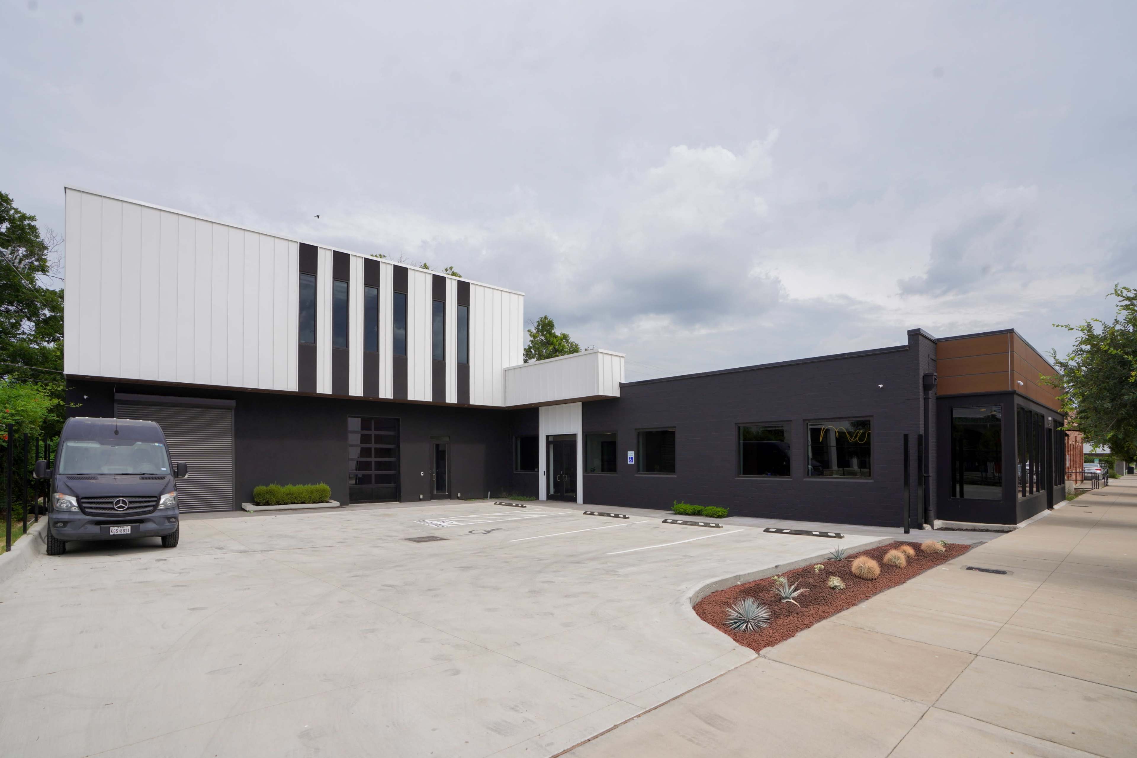 The image shows a modern building with a combination of black and white facades, featuring large windows and a paved parking area.