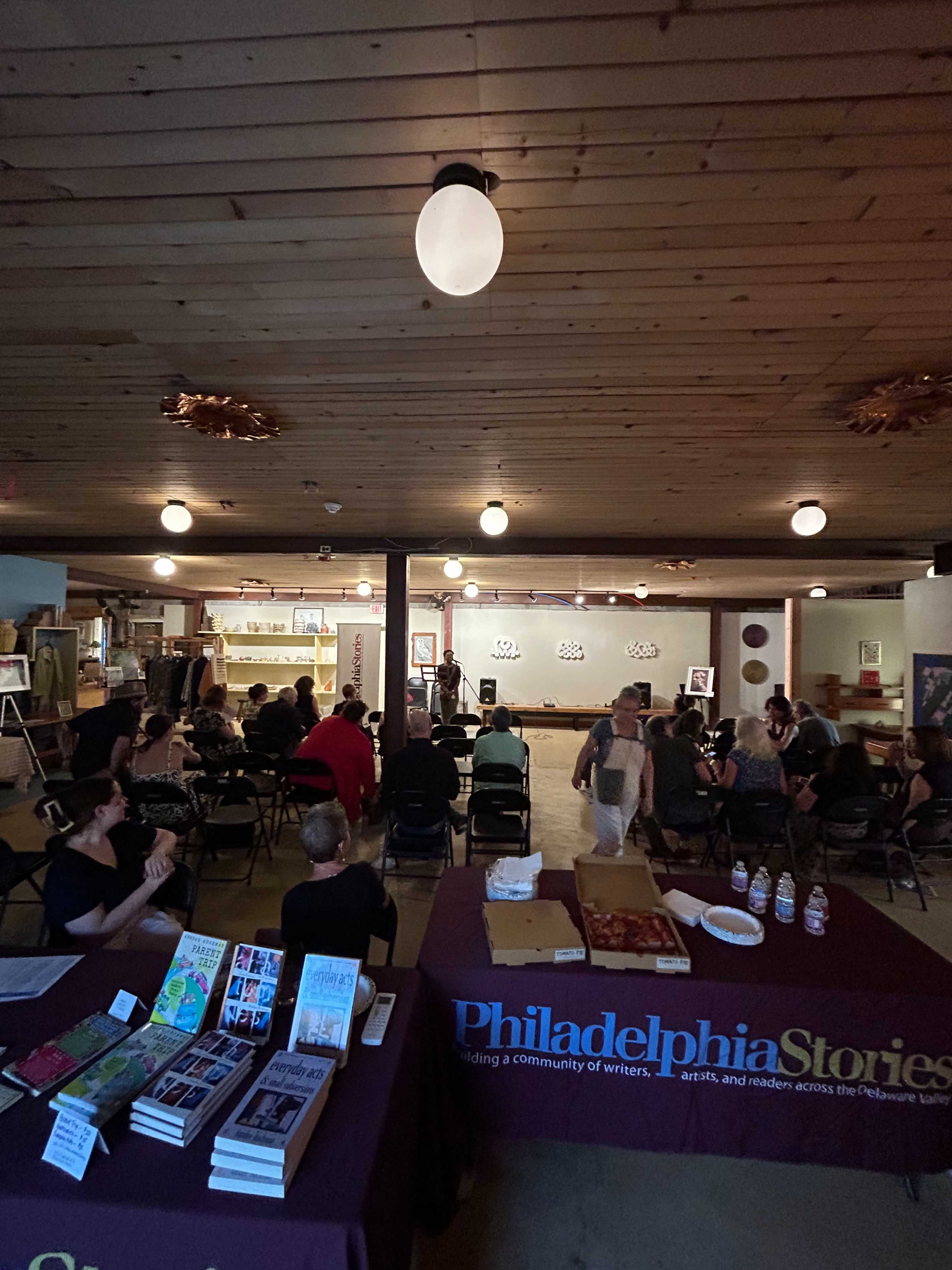A group of people sits in chairs facing an art display in a community space, with a table featuring books and snacks in the foreground.