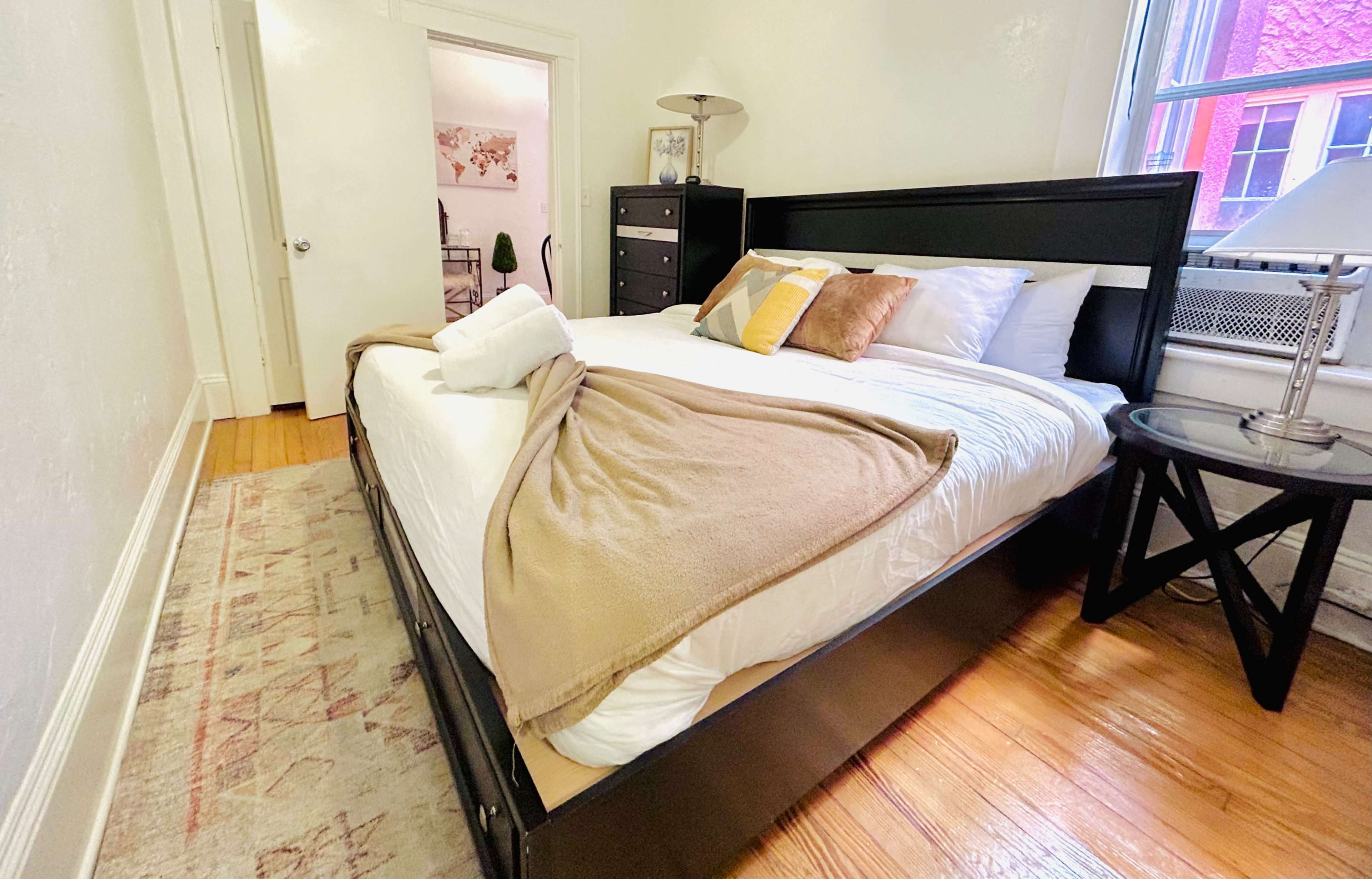 A neatly arranged bedroom features a black bed with white bedding and a light brown blanket, alongside a bedside table and a large window.