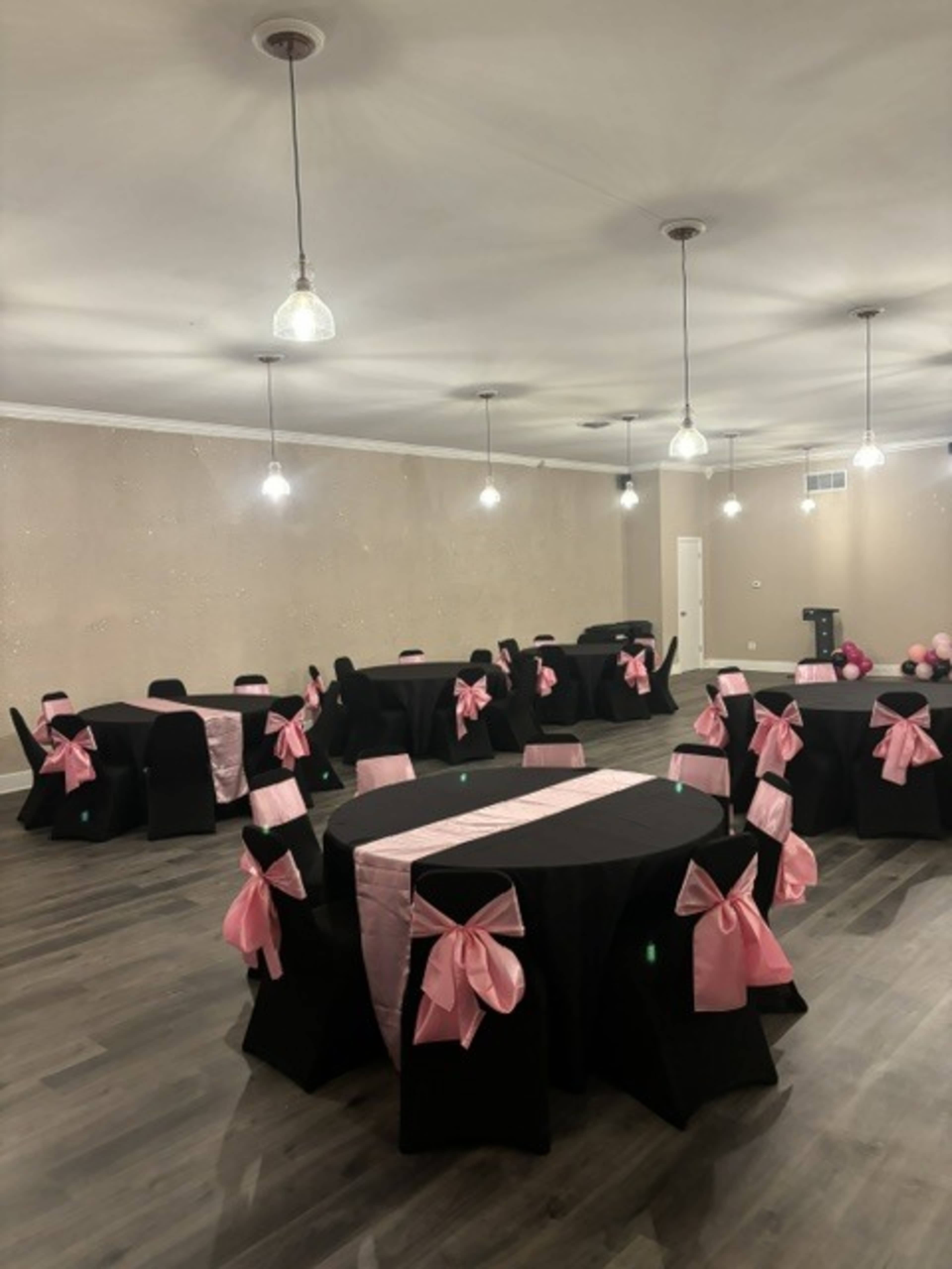 The image shows a decorated event space with several round tables covered in black tablecloths and adorned with pink bows on the chairs.