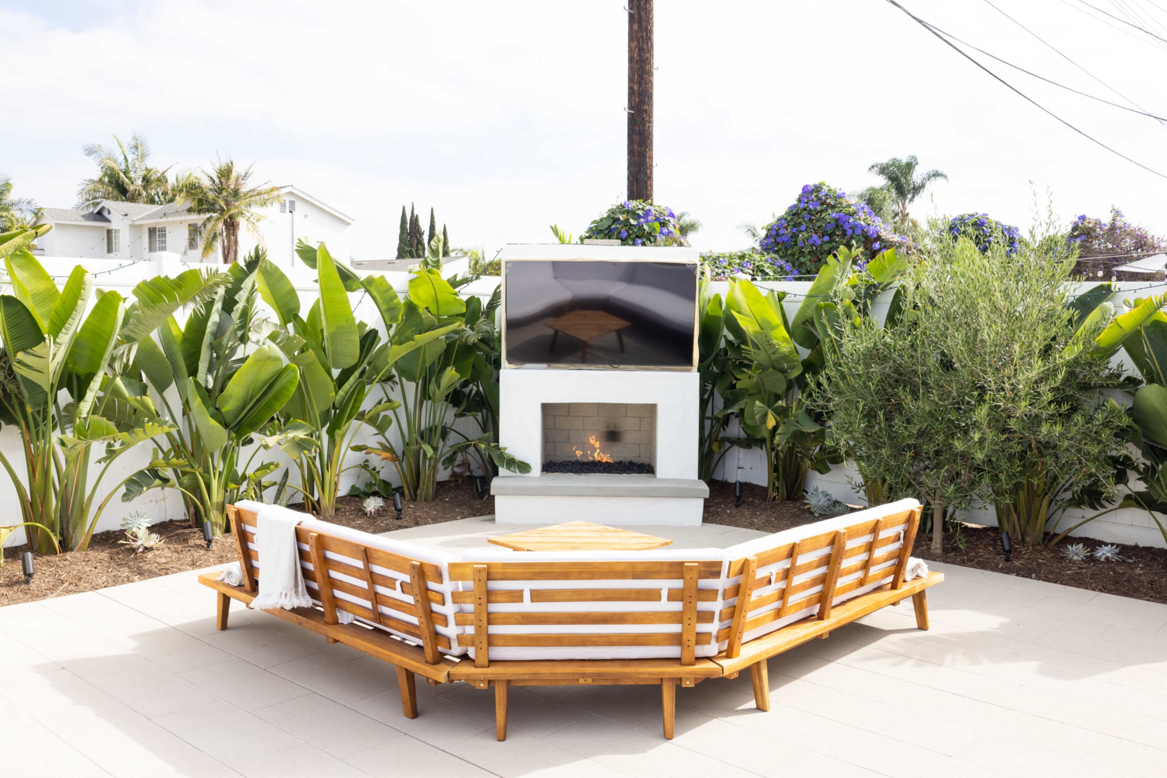 A modern outdoor seating area features a circular wooden couch around a fireplace, with a mounted TV and lush green plants in the background.