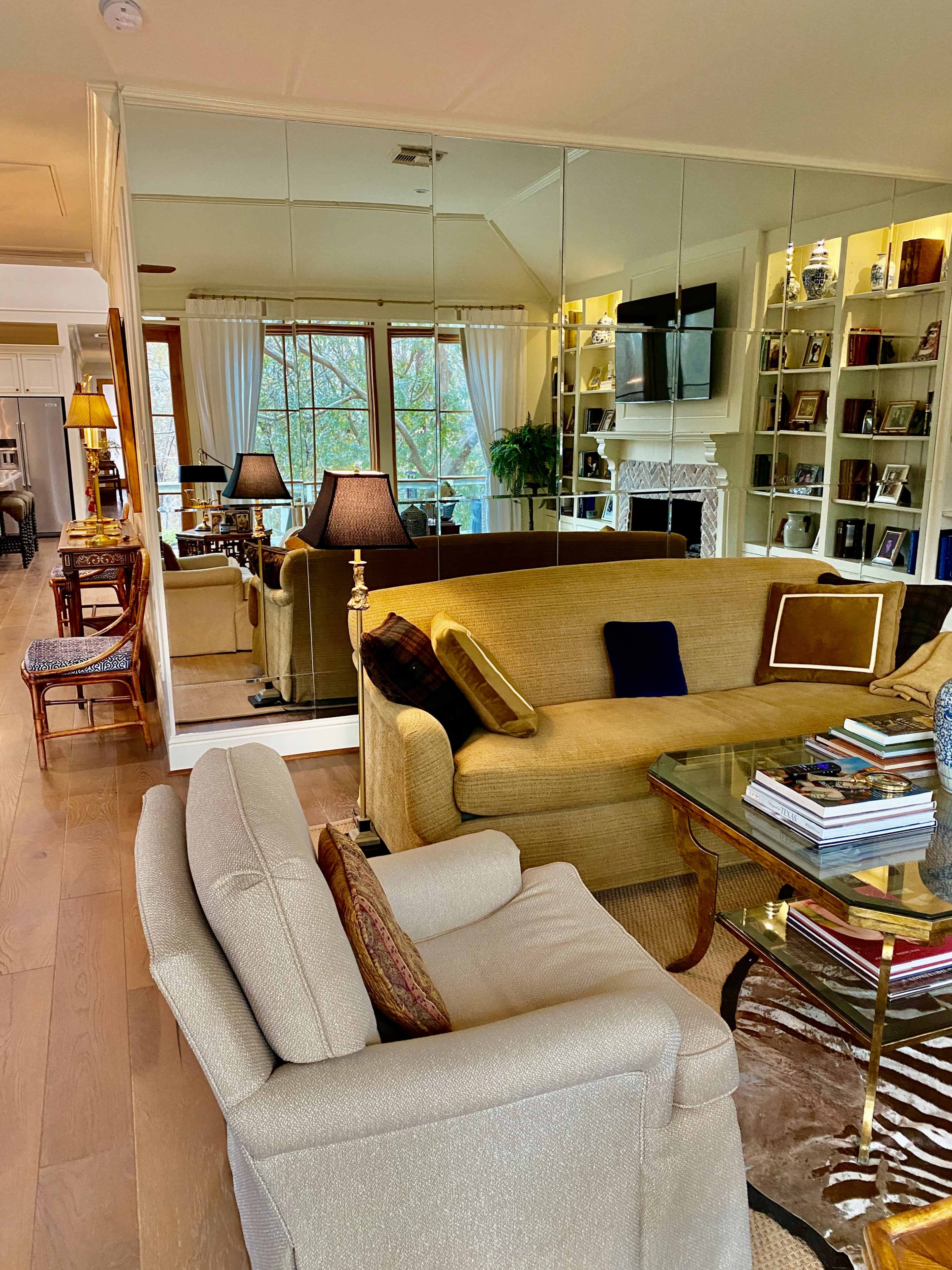 A spacious living room features a mirrored wall, a yellow couch, a beige armchair, and built-in shelves filled with books, all illuminated by natural light.