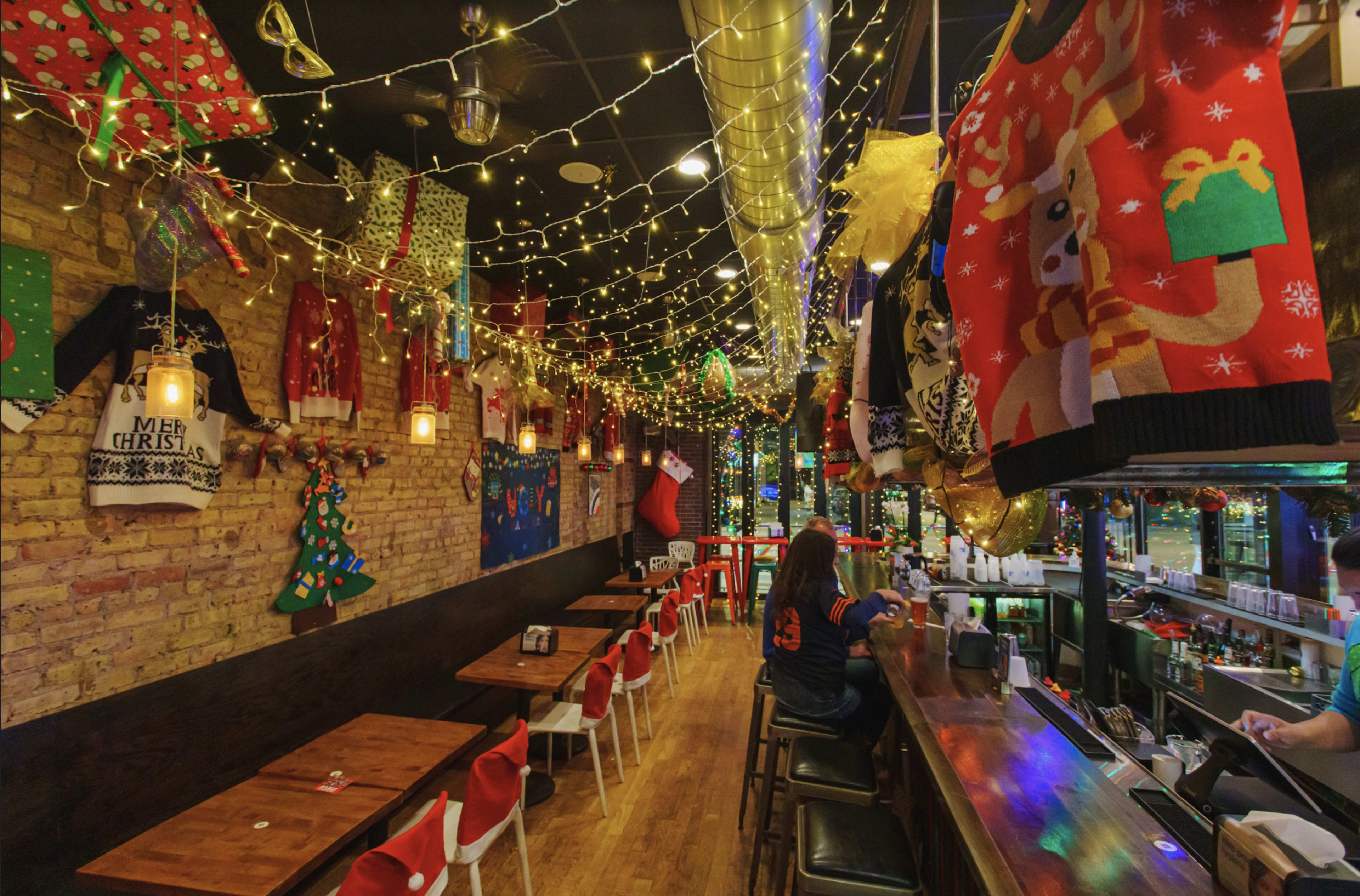 The interior of a festive restaurant features decorated walls with Christmas sweaters and lights, as patrons sit at wooden tables near a bar.
