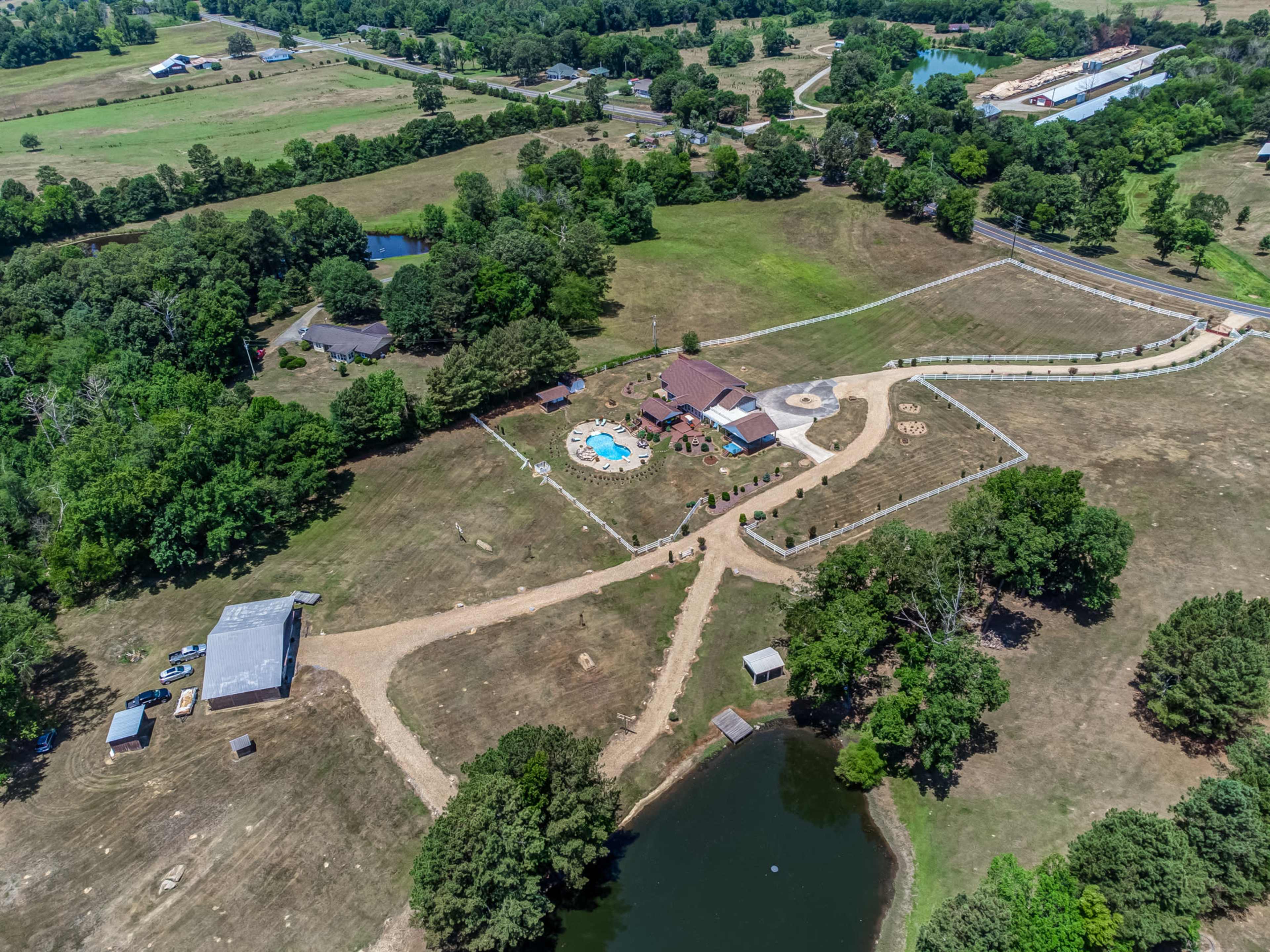 The image shows an aerial view of a rural property featuring a house with a pool, surrounded by landscaped grounds, a pond, and multiple outbuildings.