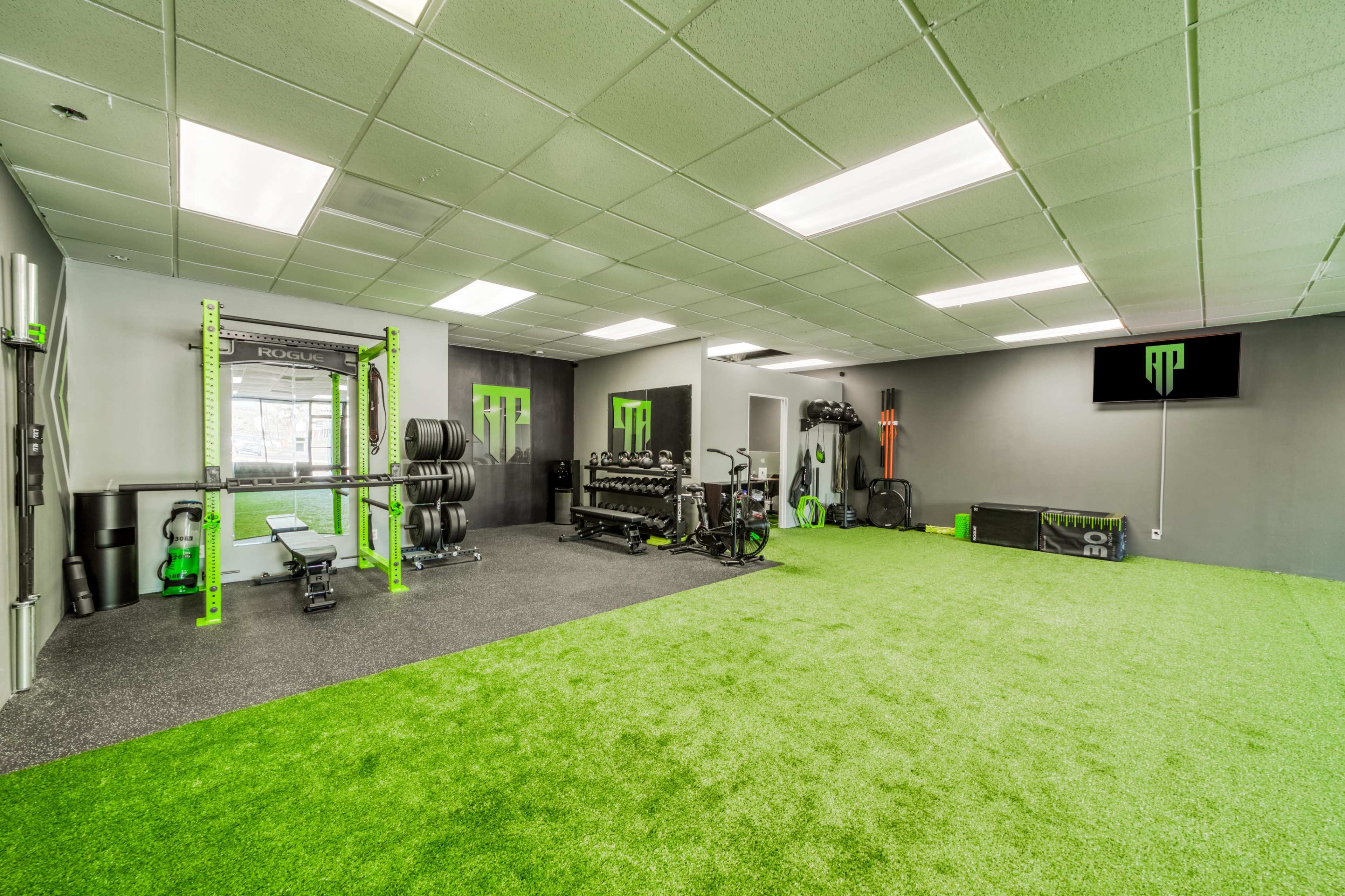 A well-equipped fitness studio featuring green turf flooring, weightlifting equipment, and cardio machines.