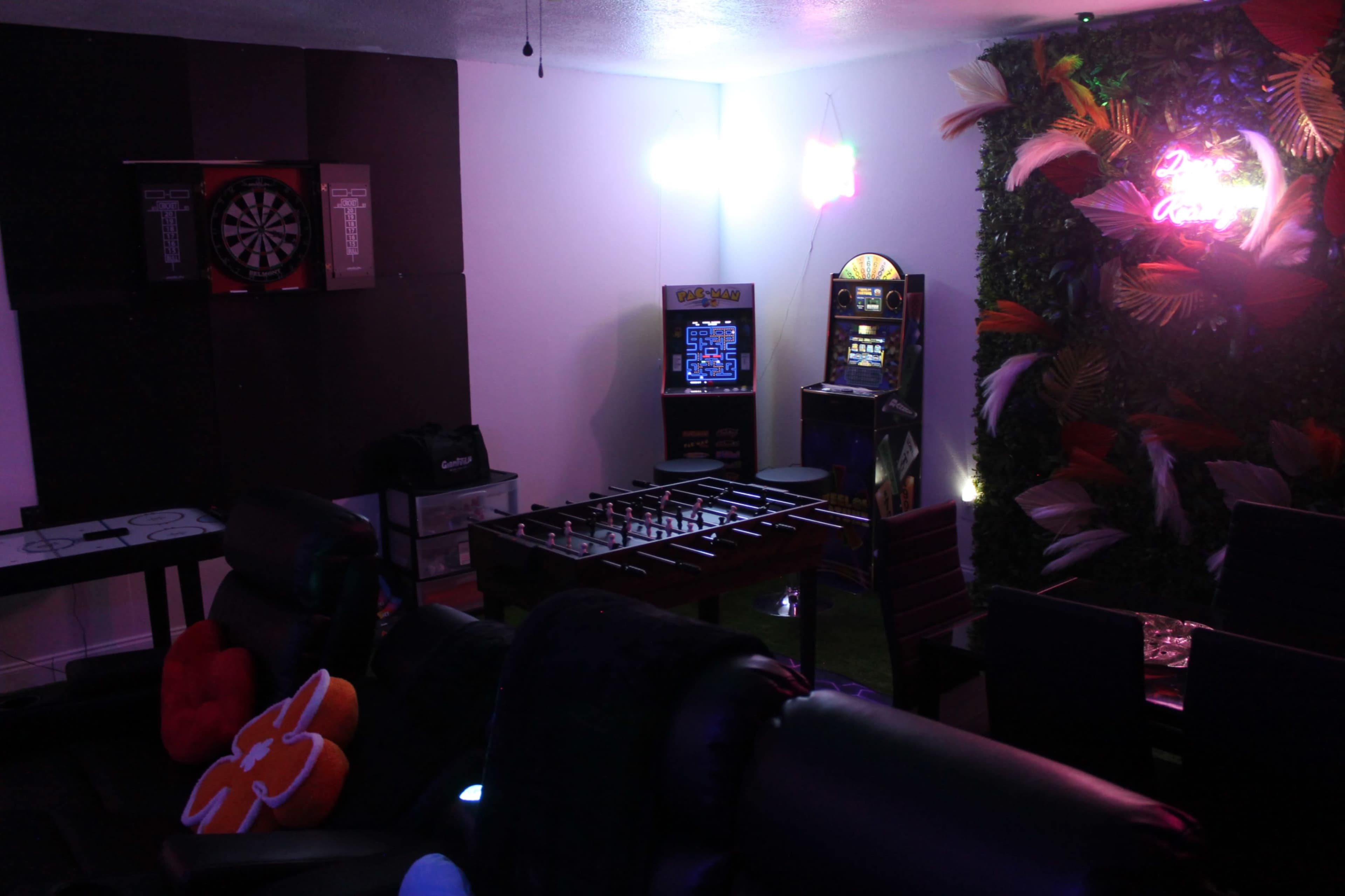 A game room featuring a dartboard, an arcade machine, a foosball table, and various seating options against a colorful wall with decorative plants.