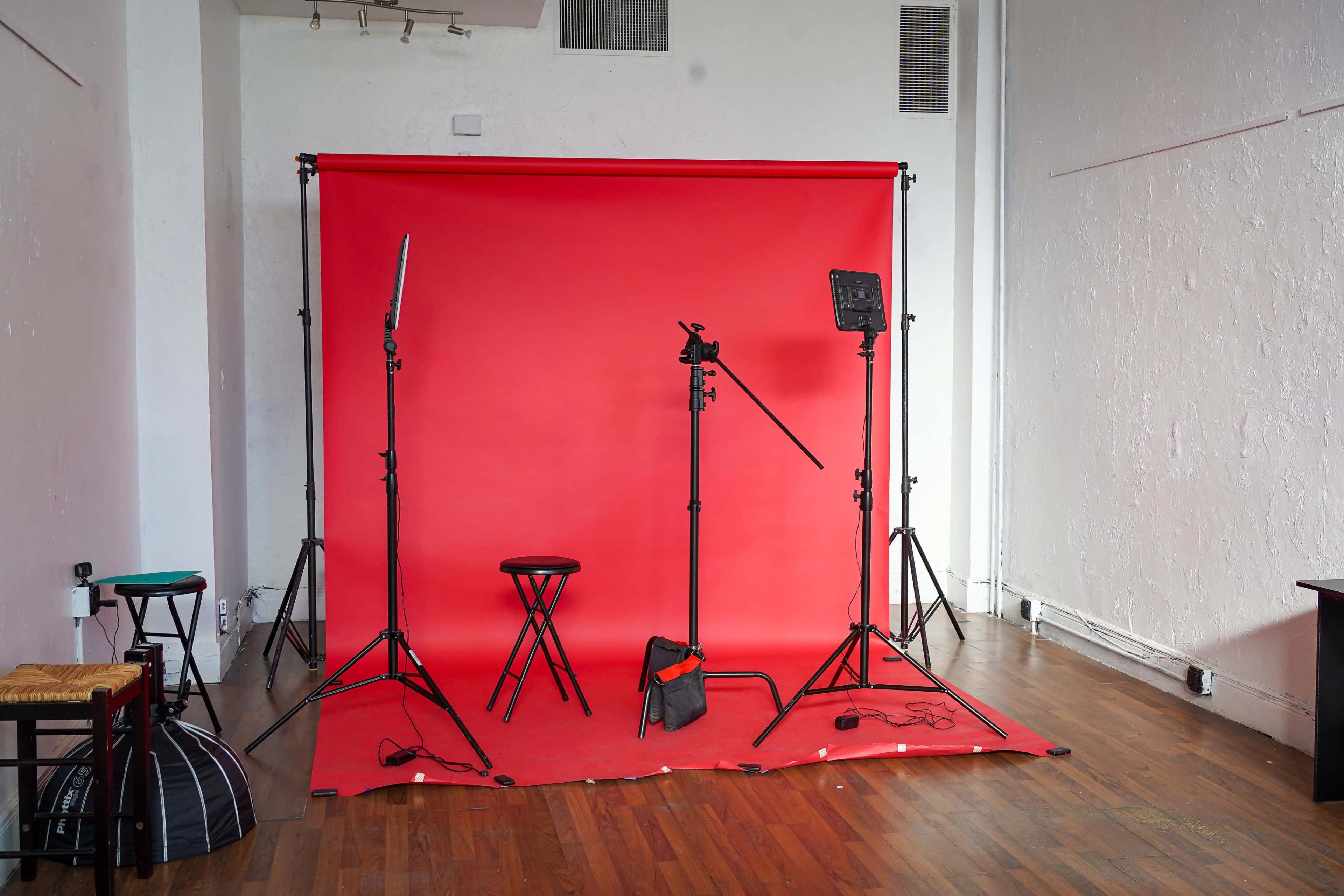 A photo studio setup with a red backdrop, three light stands, a microphone stand, a stool, and equipment on the wooden floor.