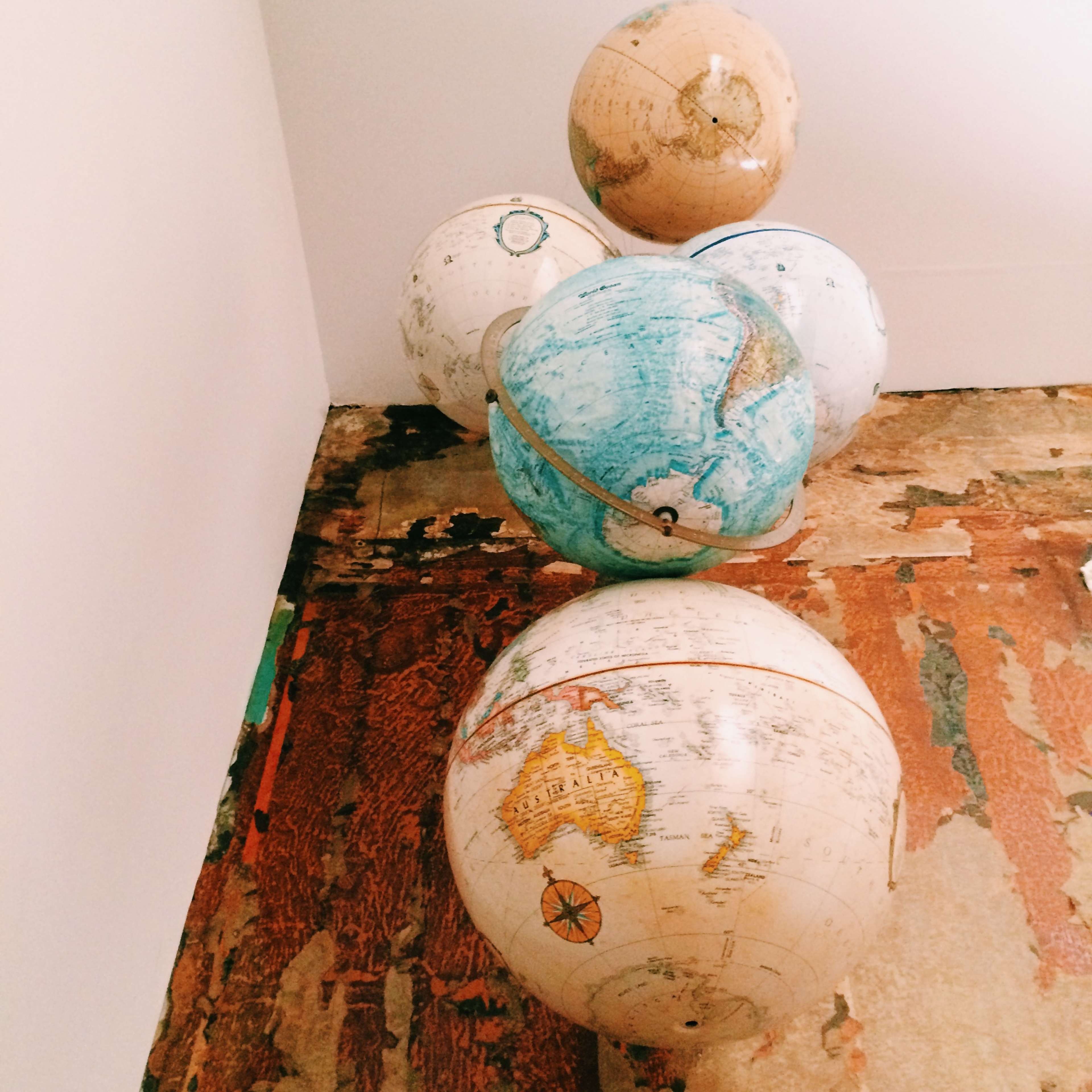 Five vintage globes are stacked in a corner on a wooden floor.