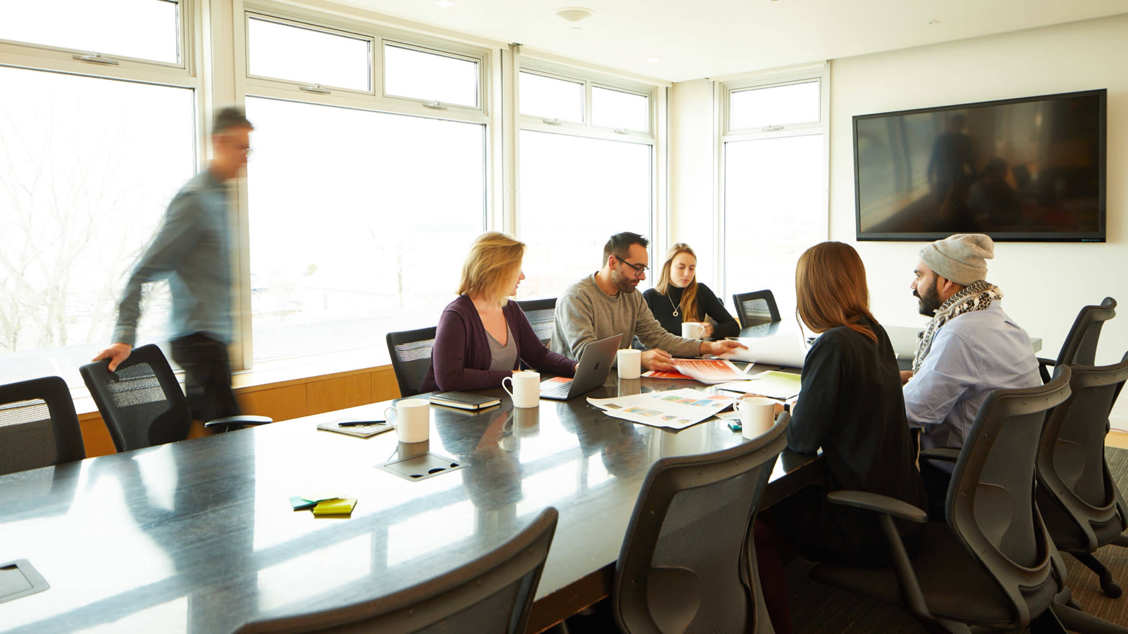 A group of six people are seated around a long conference table in a well-lit meeting room, discussing documents while one person walks past.
