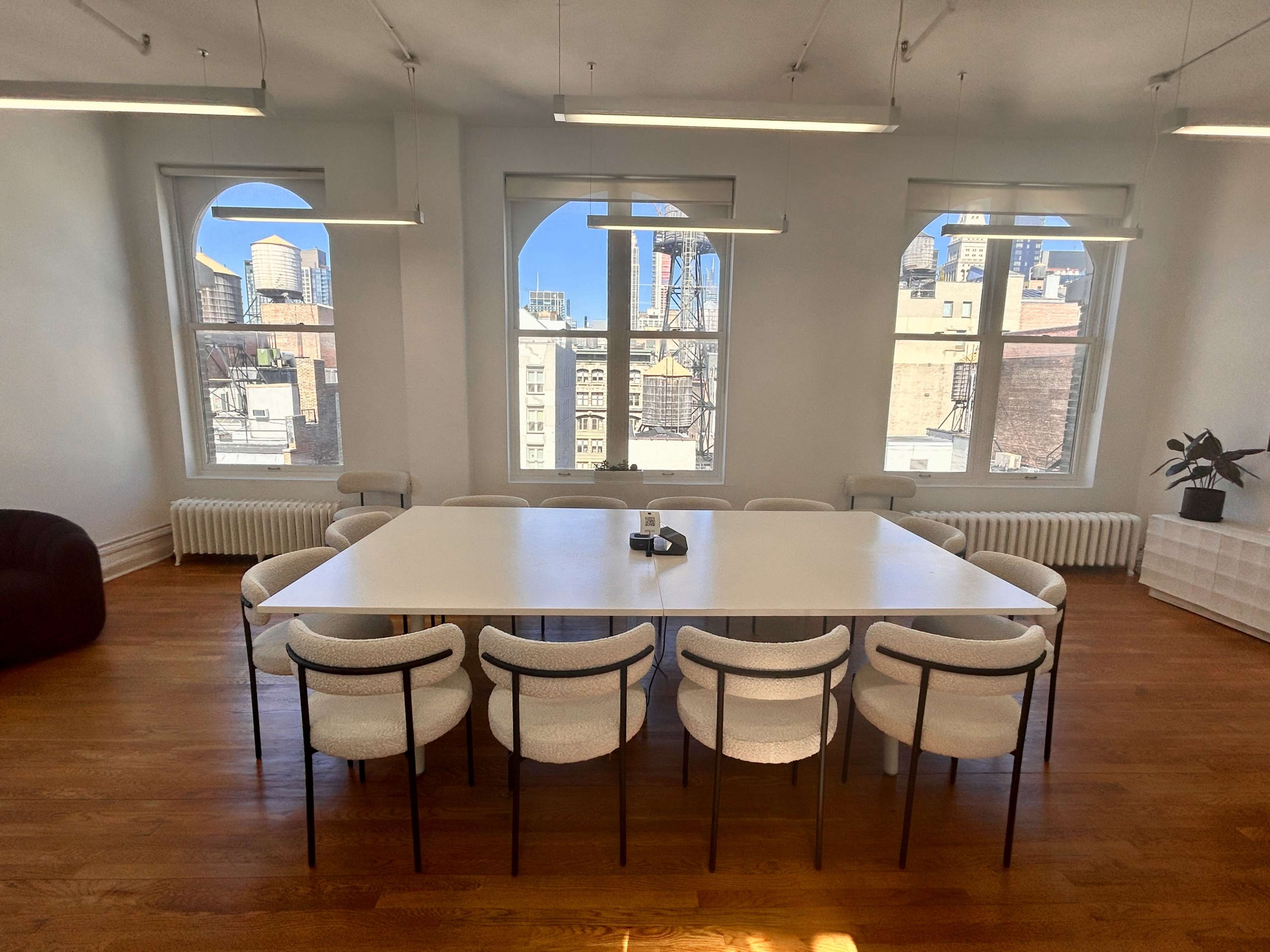 A conference room with a large white table surrounded by ten chairs, featuring three tall windows that overlook a city skyline.