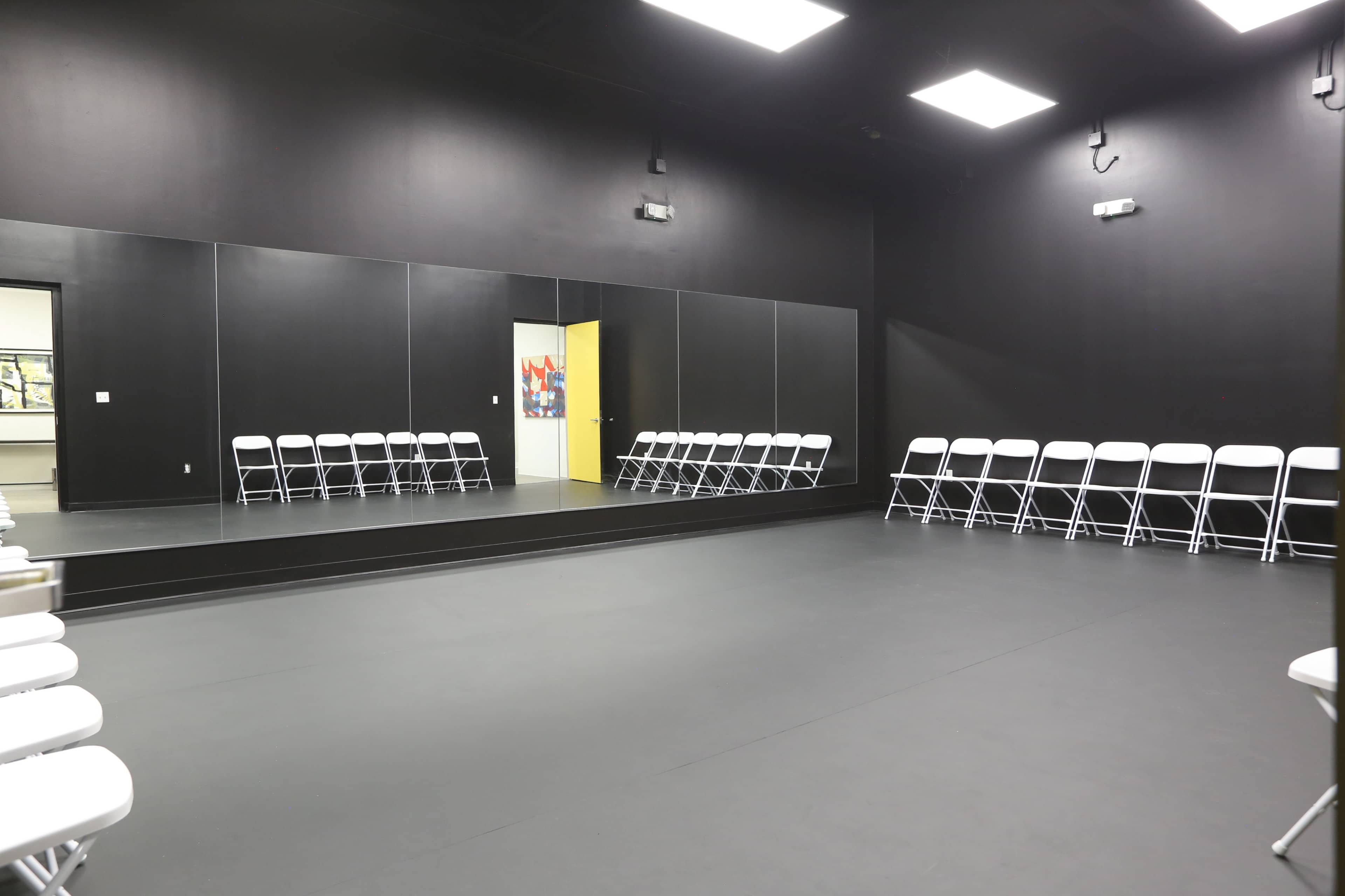 A large, empty room with black walls, a mirrored wall, and several white folding chairs arranged neatly against one side.
