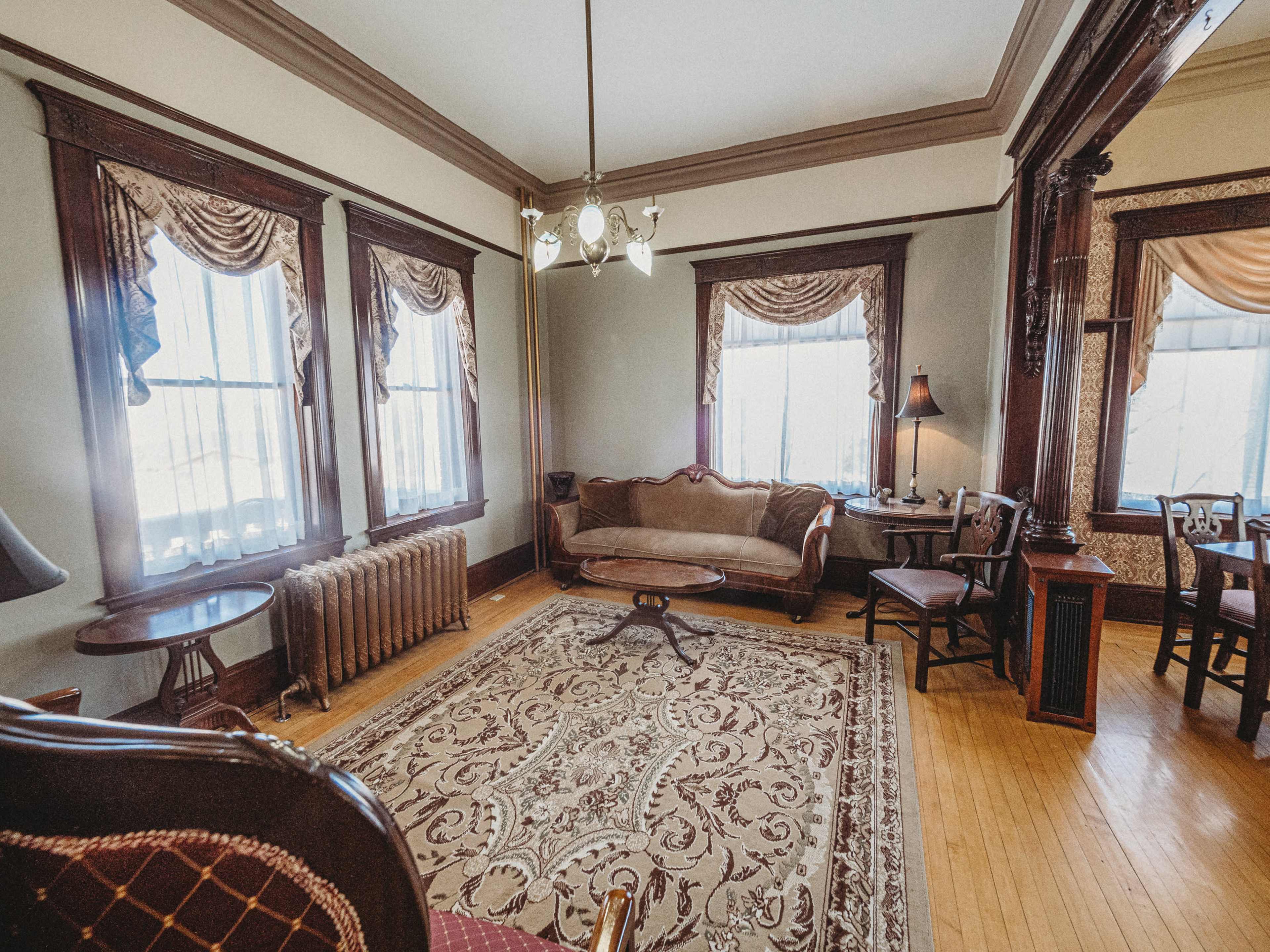 The scene shows a warmly lit living room with hardwood floors, ornate furniture, and large windows draped with curtains, featuring a central area rug.