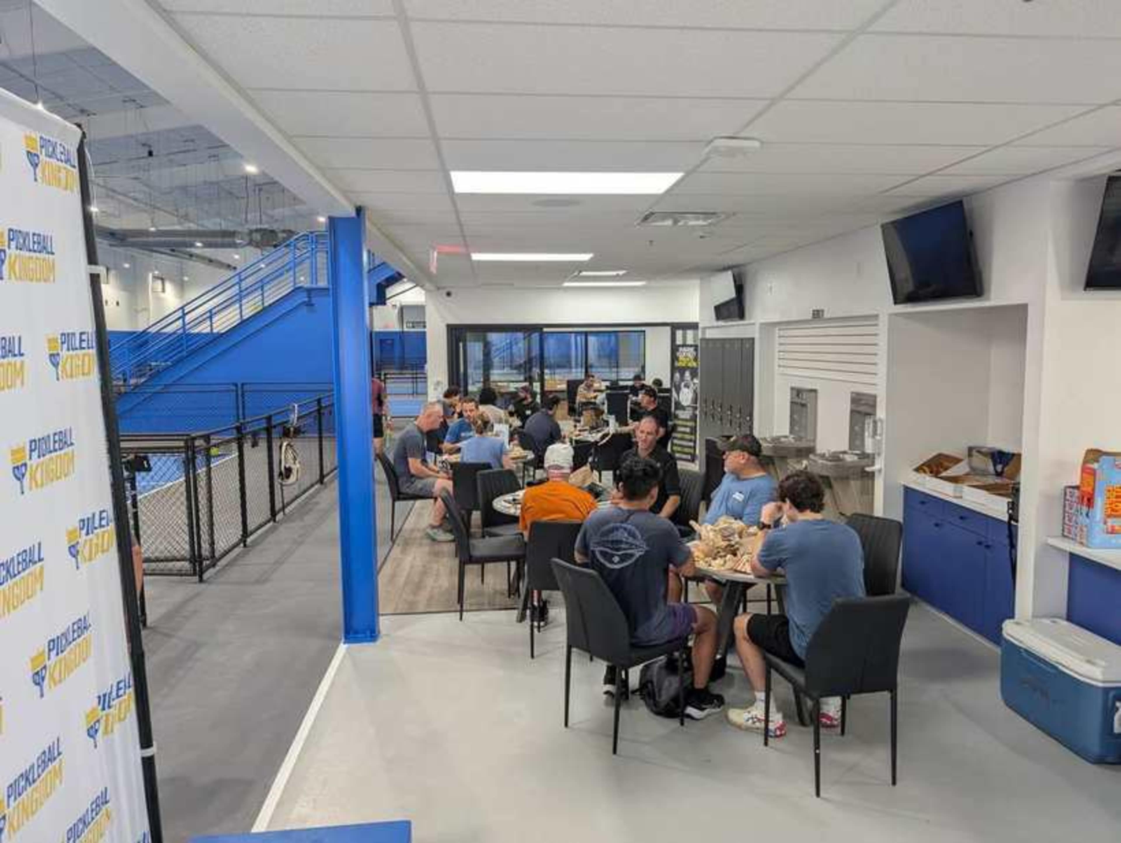 A group of people is seated at tables enjoying a meal in a spacious cafeteria with blue and white decor.