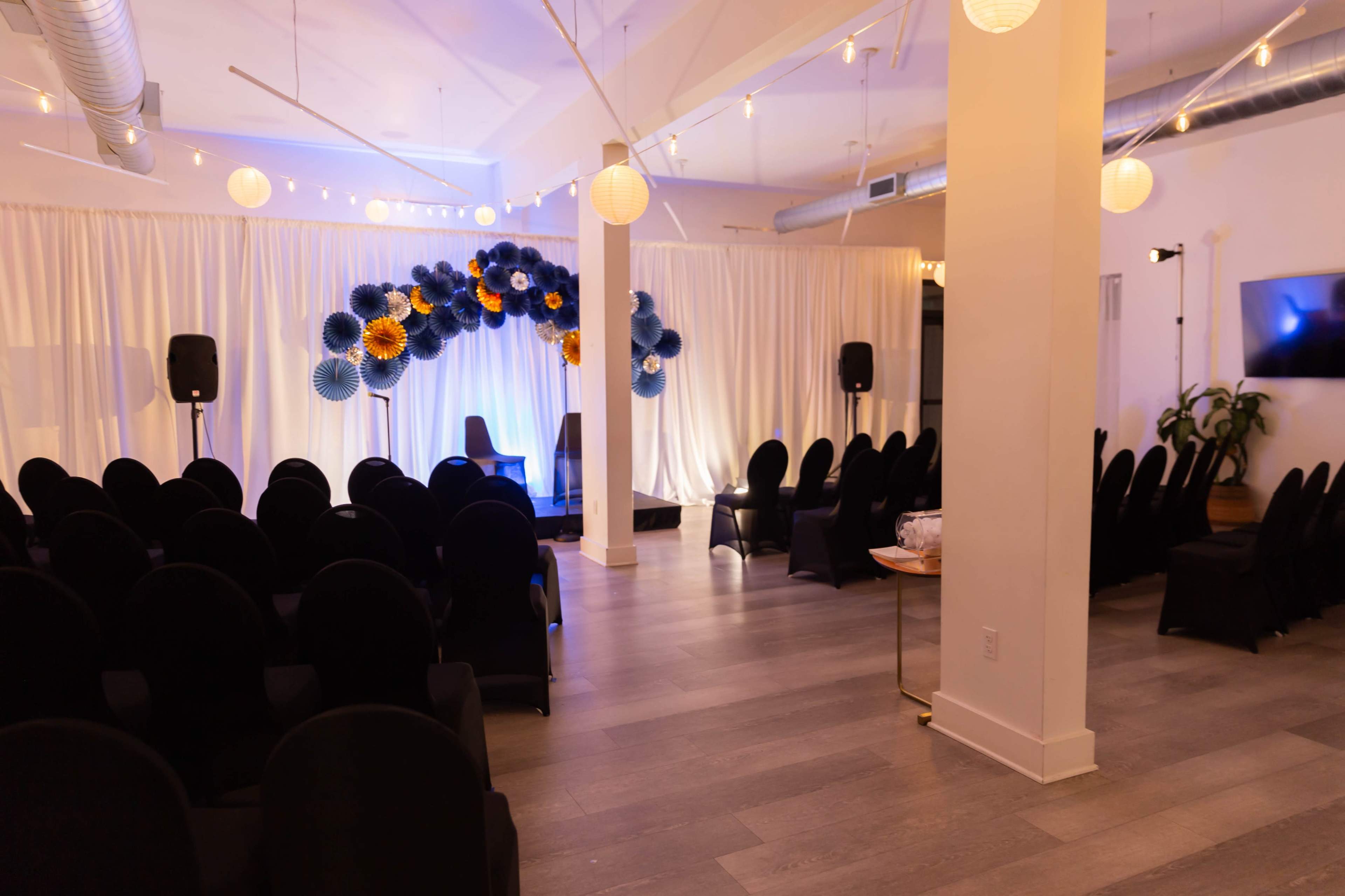 The image shows an event space set up with black chairs facing a stage decorated with colorful paper decorations and curtains.