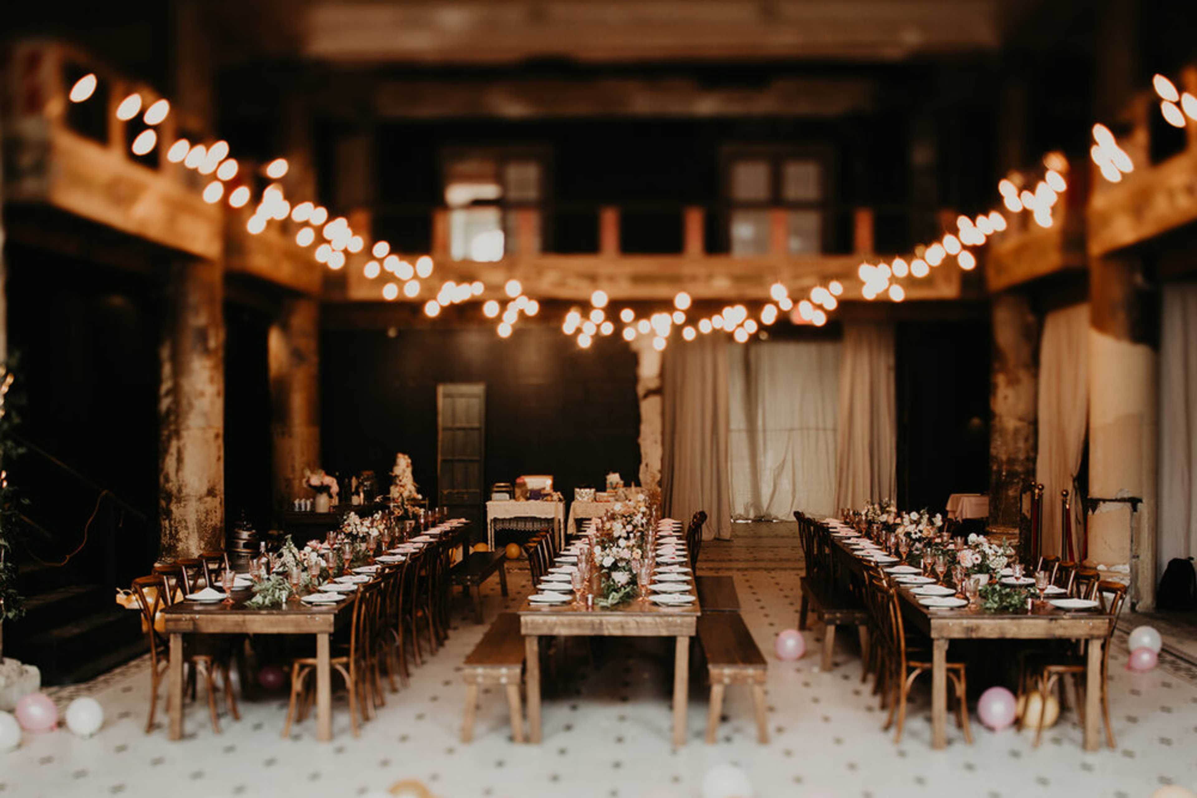 A long banquet hall features multiple wooden tables set for dining, adorned with floral centerpieces and string lights overhead.