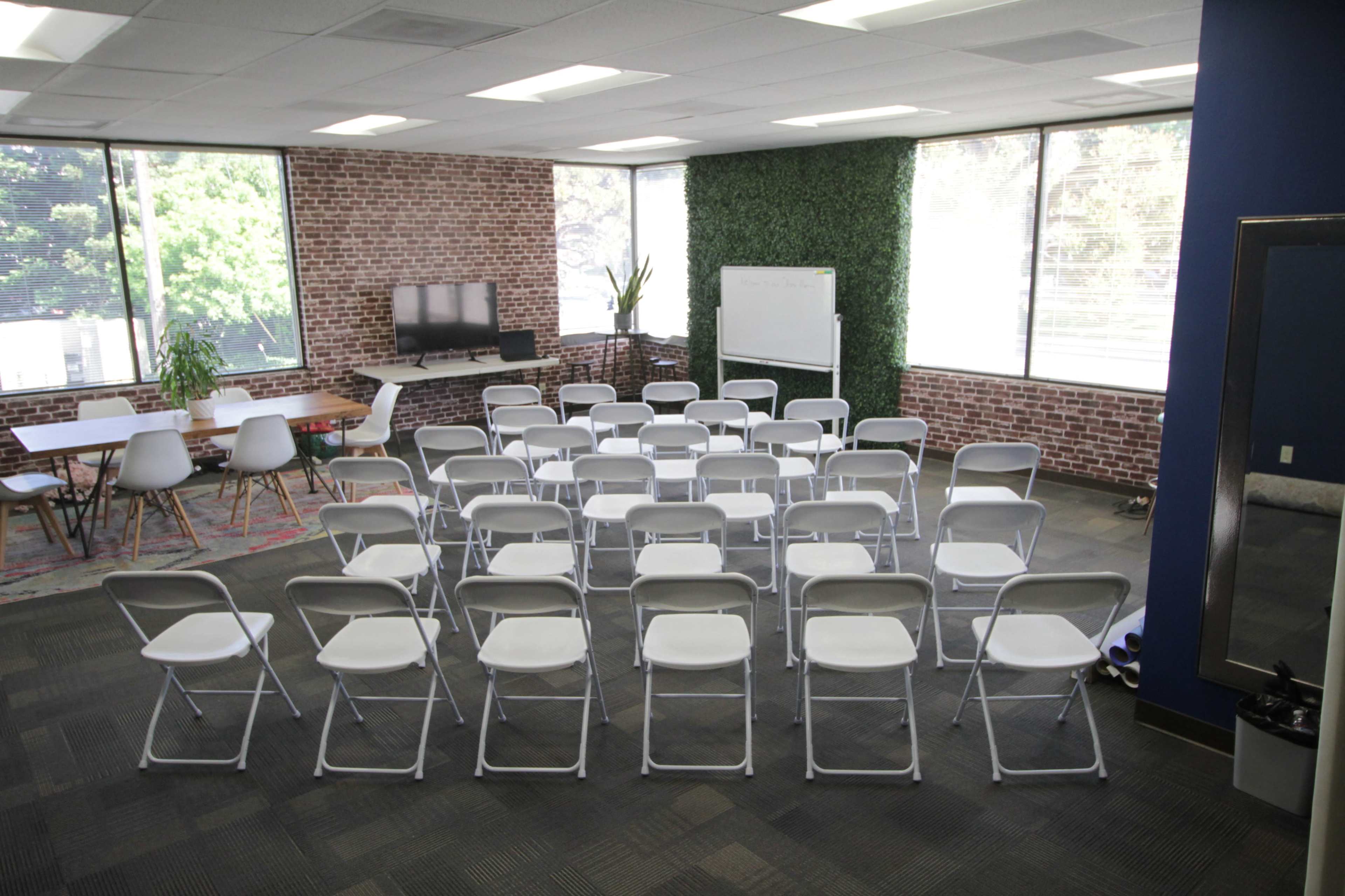 A meeting room features a layout of white folding chairs arranged in rows, with a TV, a whiteboard, and greenery visible through large windows.