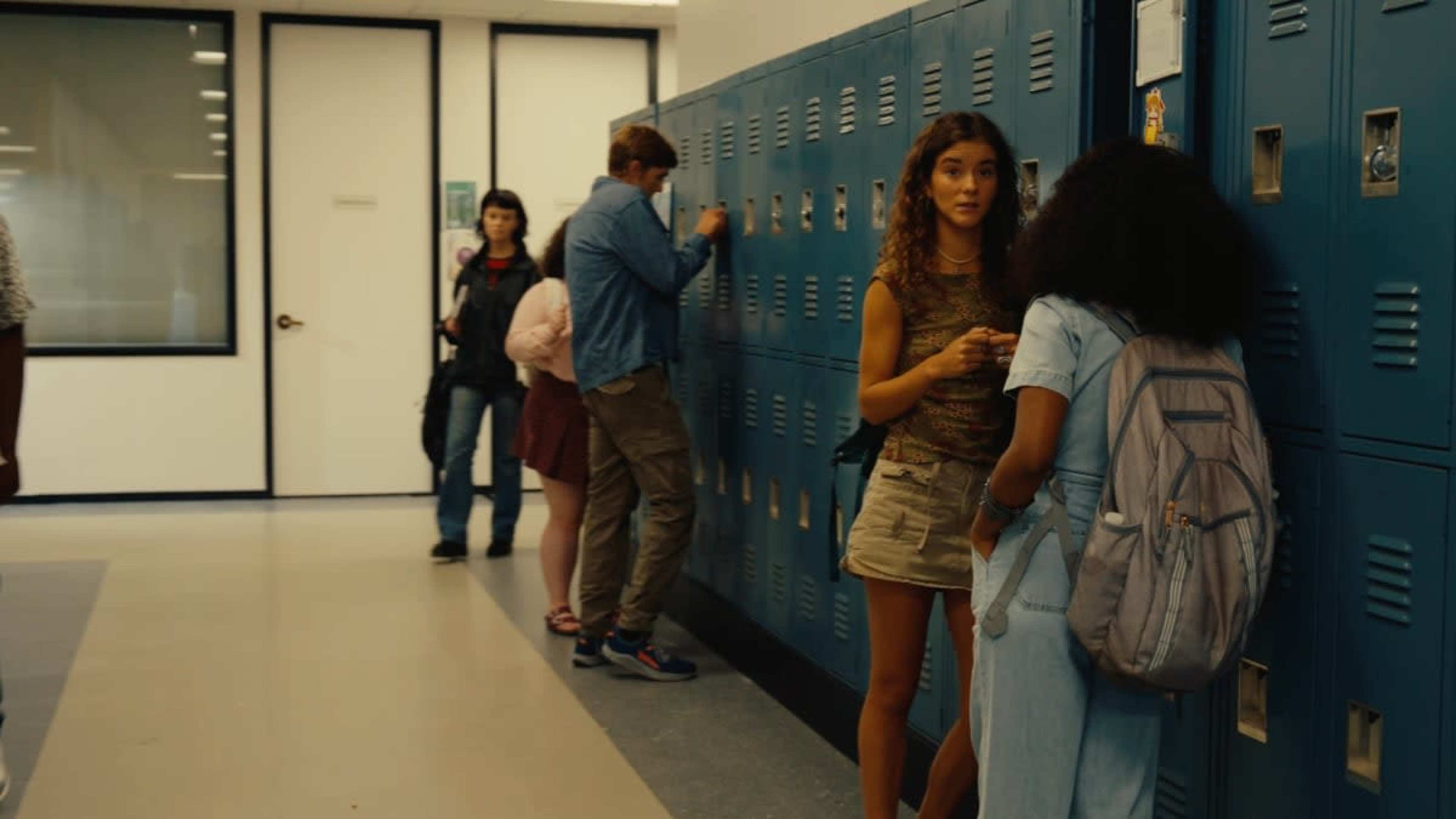 A group of students stands in a school hallway next to lockers, with two girls chatting while another student looks inside a locker.
