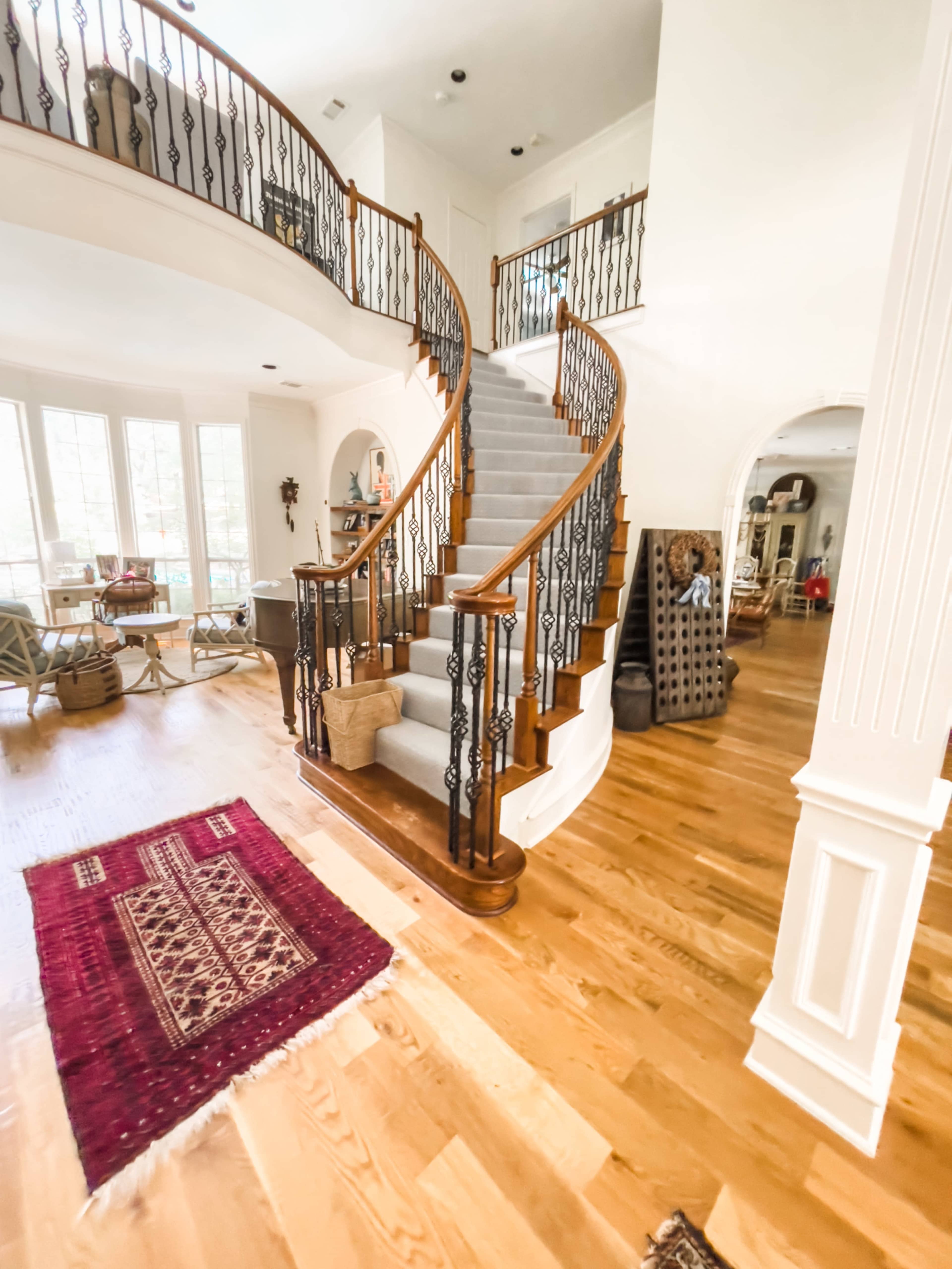 A curved wooden staircase with black wrought iron railing ascends from a spacious foyer featuring hardwood floors and a decorative area rug.