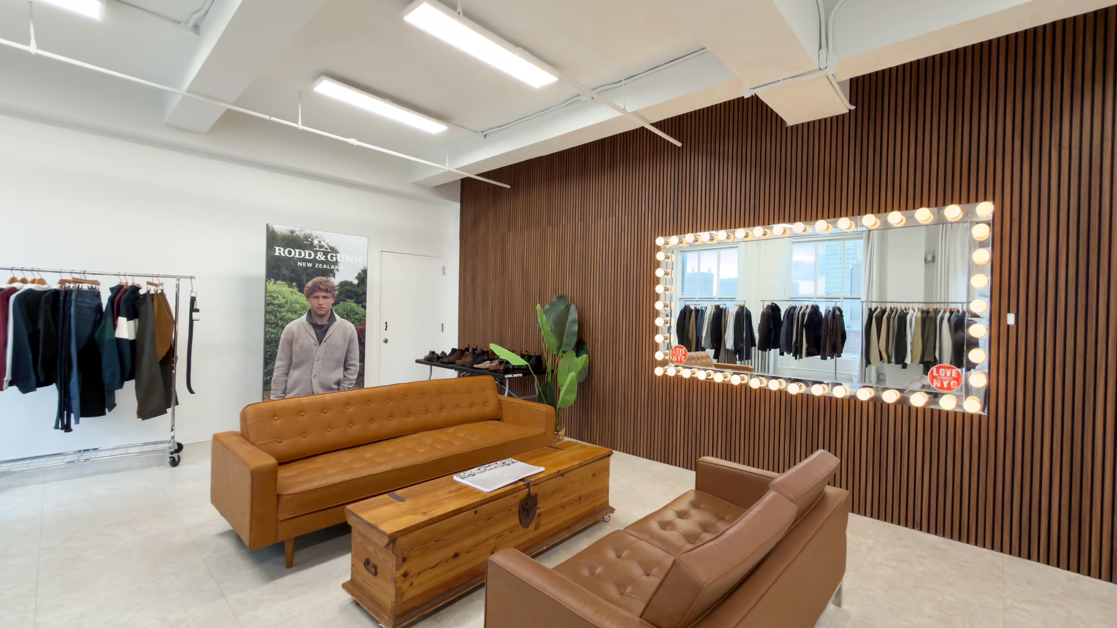 The image features a modern retail space with two brown leather sofas, a wooden coffee table, clothing racks, and a large illuminated mirror on a wooden wall.
