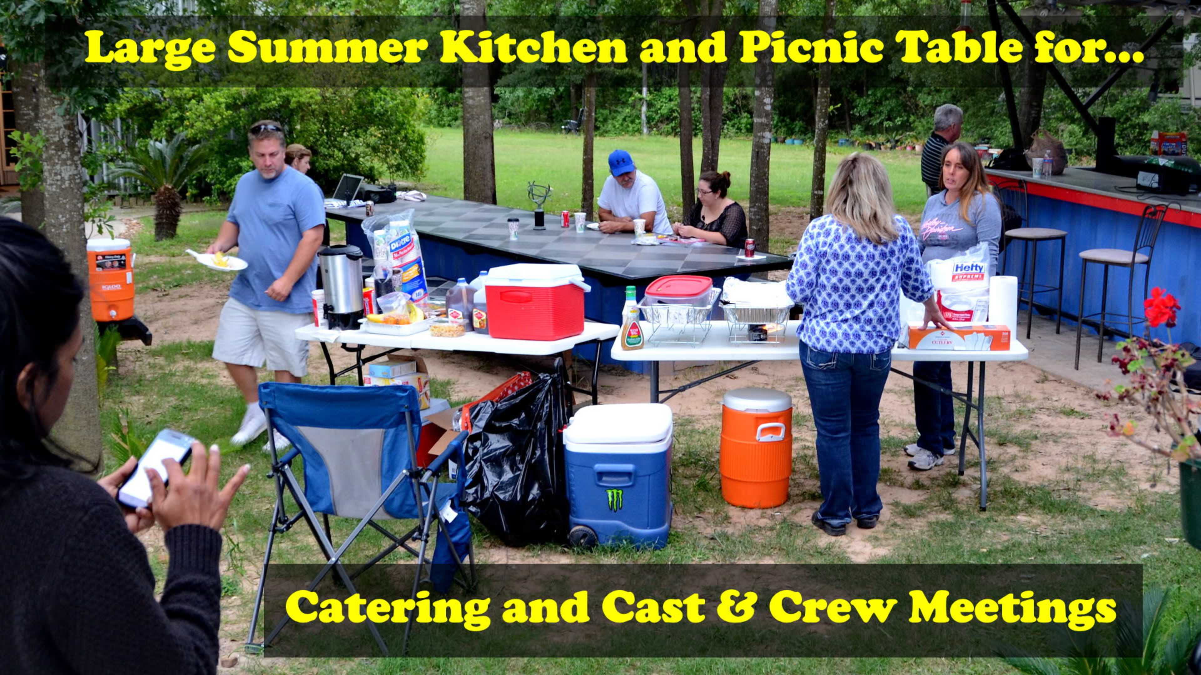 A group of people are gathered around a large outdoor kitchen and picnic table, engaging in food preparation and dining in a yard setting.