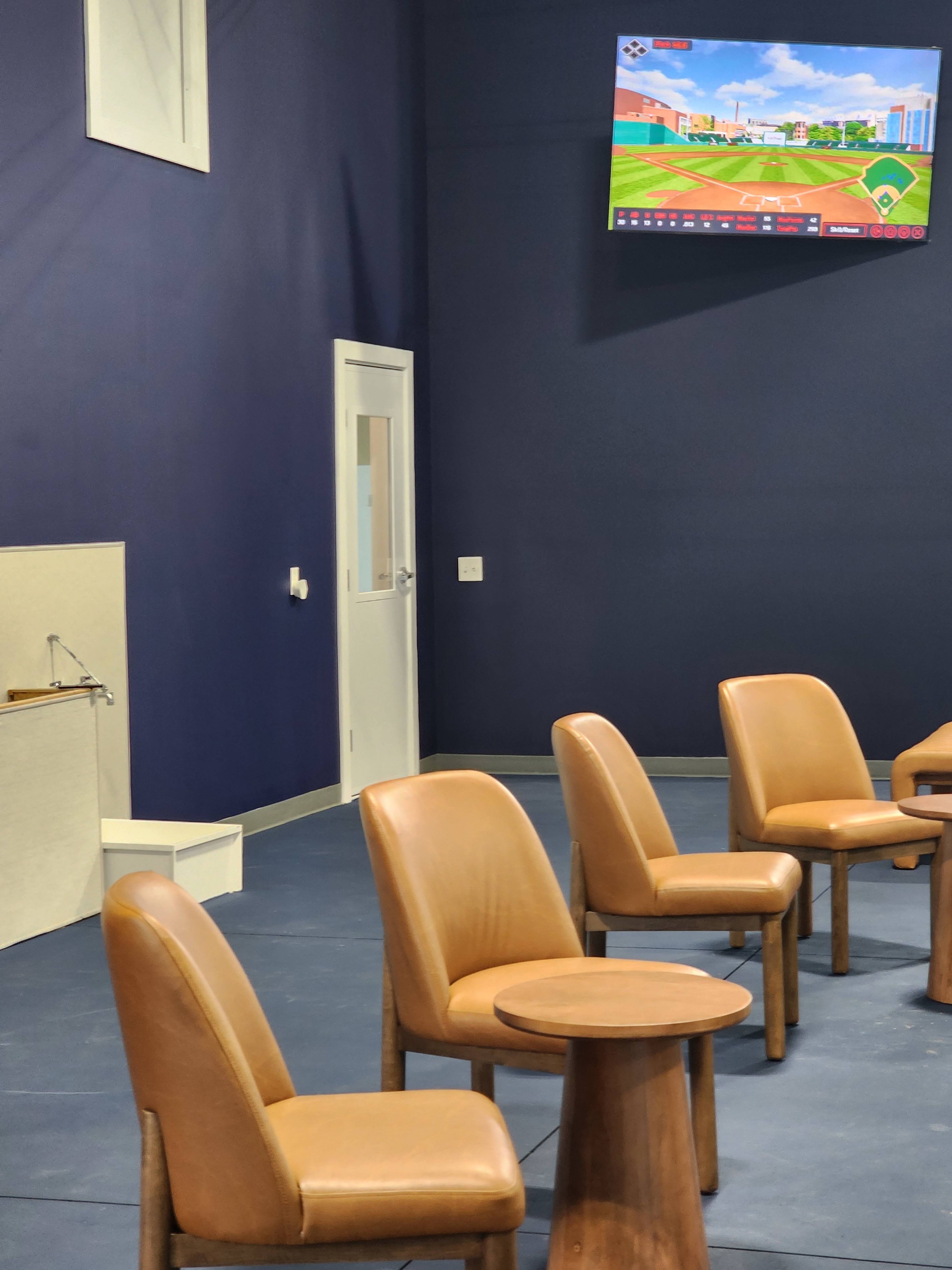 The image shows a minimalistic room with six brown chairs arranged around small wooden tables, a television mounted on the wall displaying a sports game, and a door in the background.
