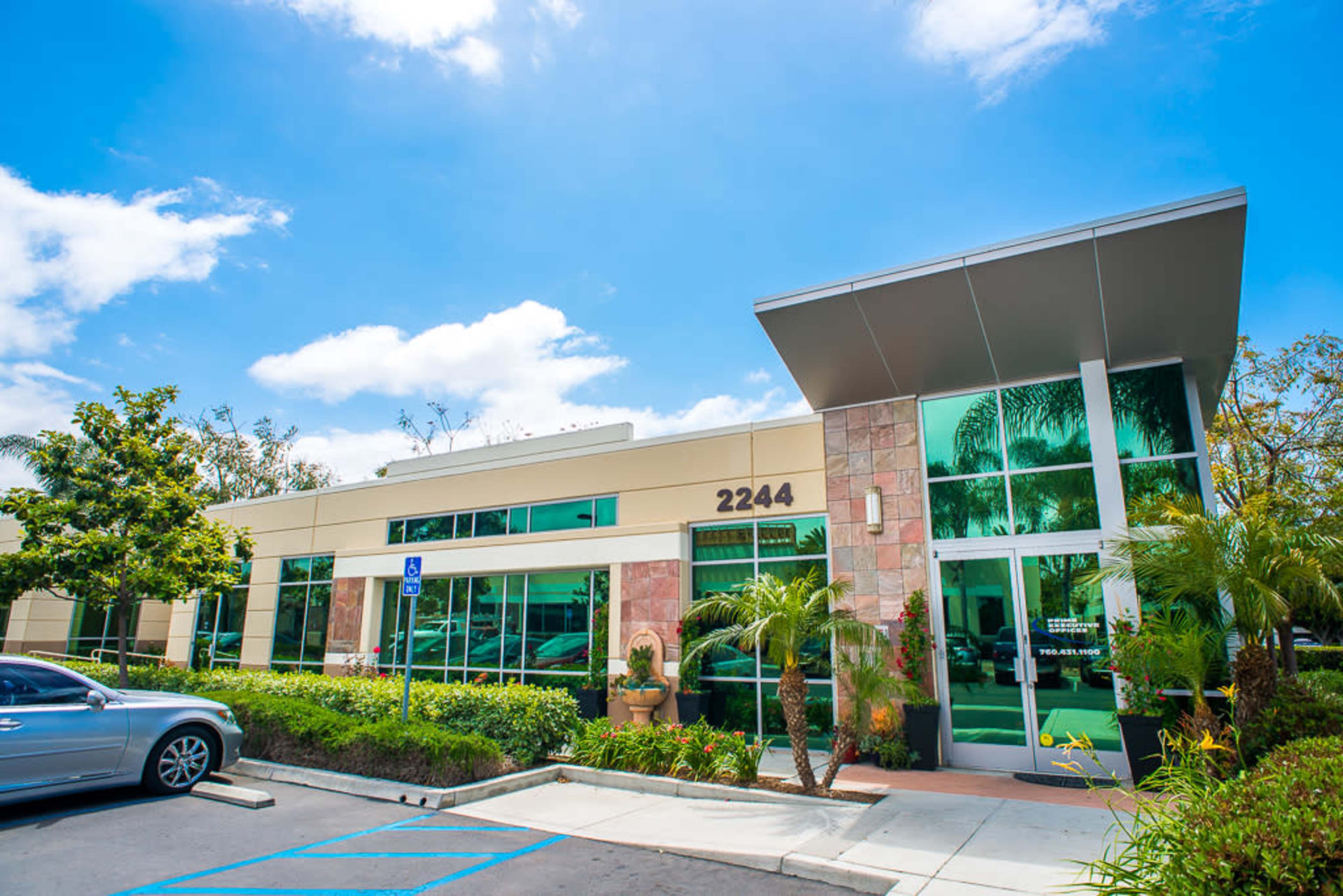 The image shows a modern commercial building with large glass windows and a stone facade, set against a bright blue sky and surrounded by greenery.