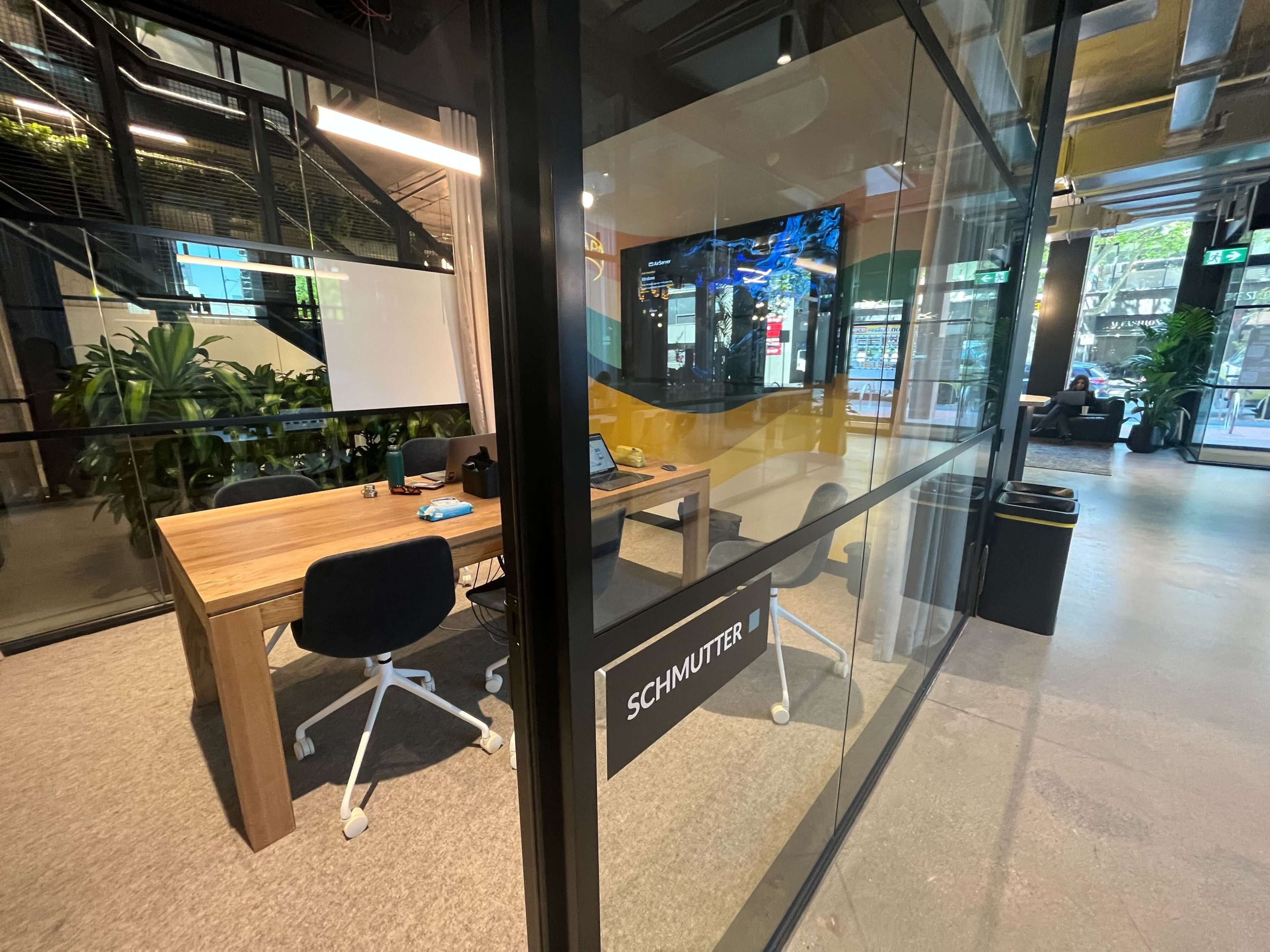 The image shows a modern office space with a glass meeting room featuring a wooden table and chairs, surrounded by plants and natural light.