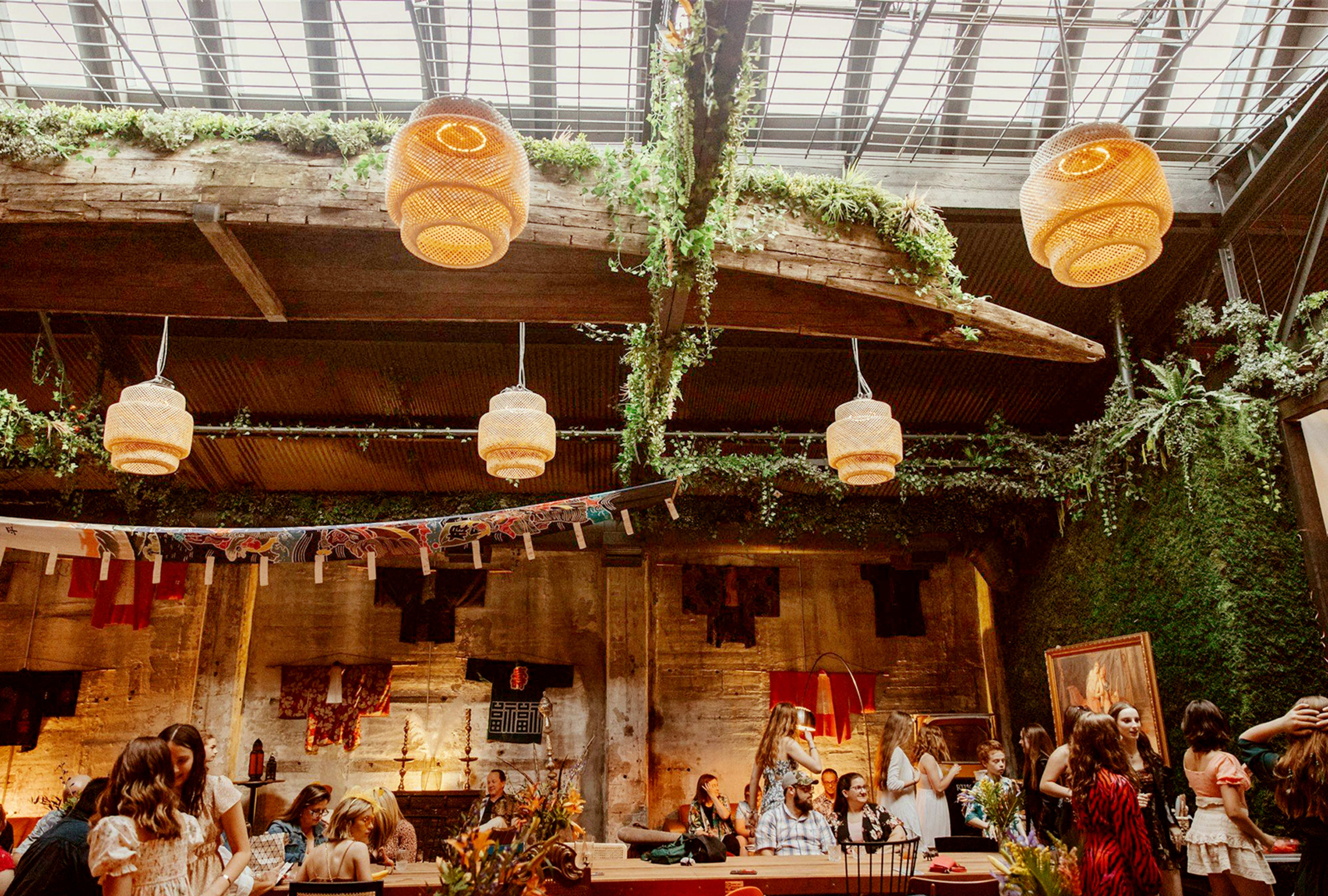 The image features a spacious rustic interior with hanging woven lights, greenery on the walls, and groups of people socializing around dining tables.