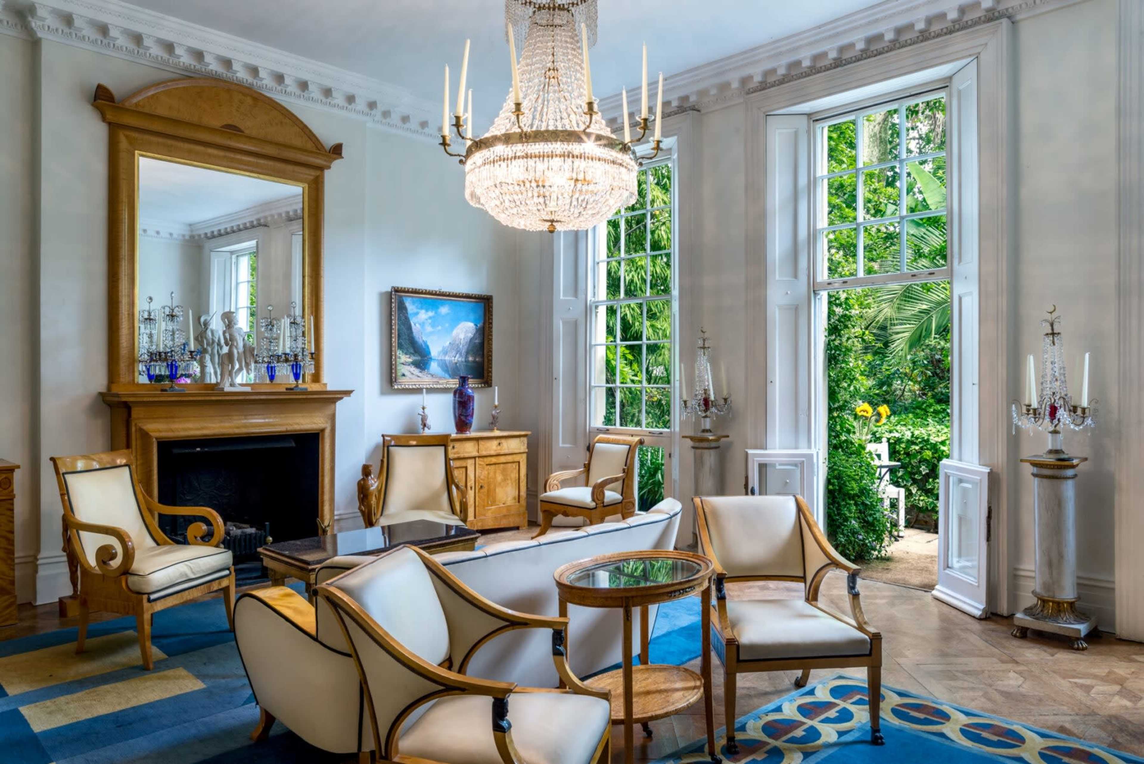 The image depicts a elegantly furnished living room featuring white chairs, a chandelier, a fireplace, and large windows that open to a garden view.