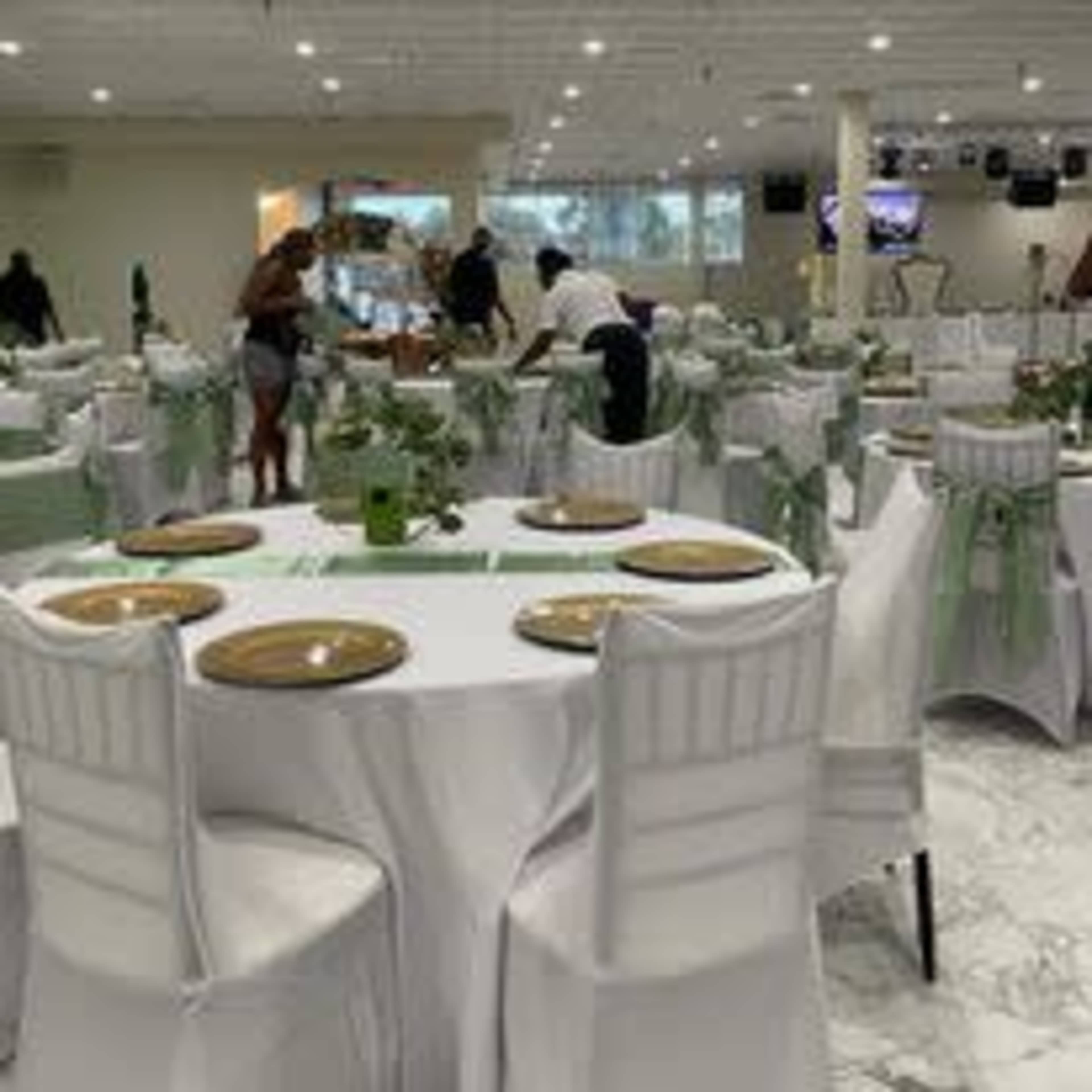 A spacious dining area is set up with white tables covered in linens, decorated with green accents and plates, as staff prepares for an event.