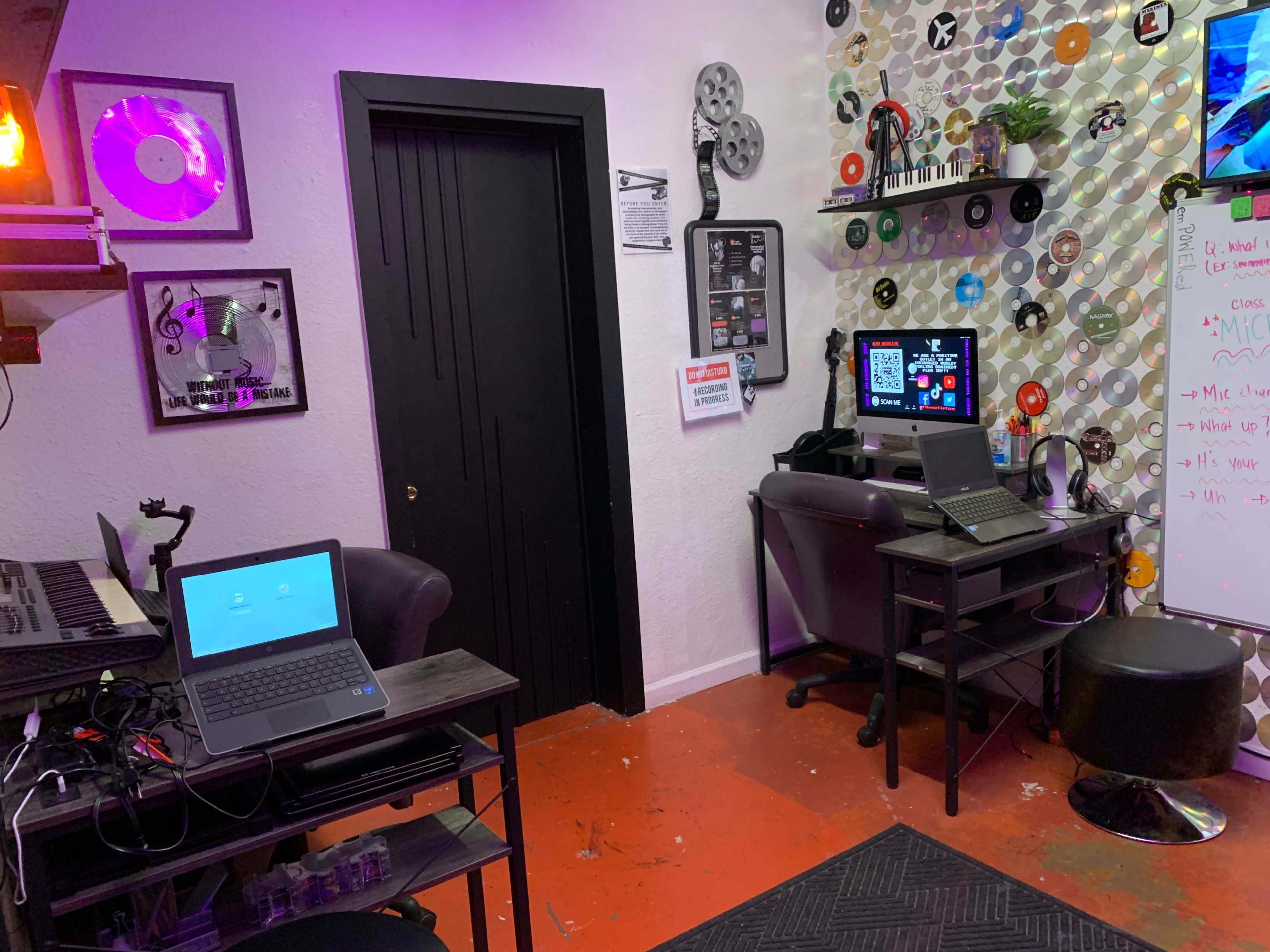 A small music studio featuring two laptops on desks, colorful wall decor made of vinyl records, and a door leading to another room.
