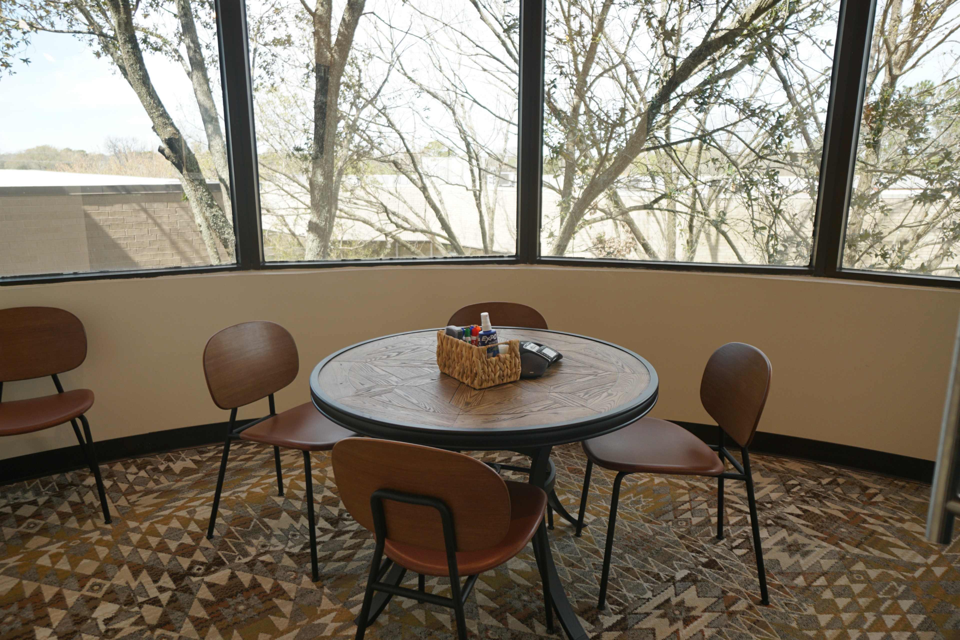 A round table with a basket of markers sits surrounded by four chairs in a room with large windows displaying trees outside.