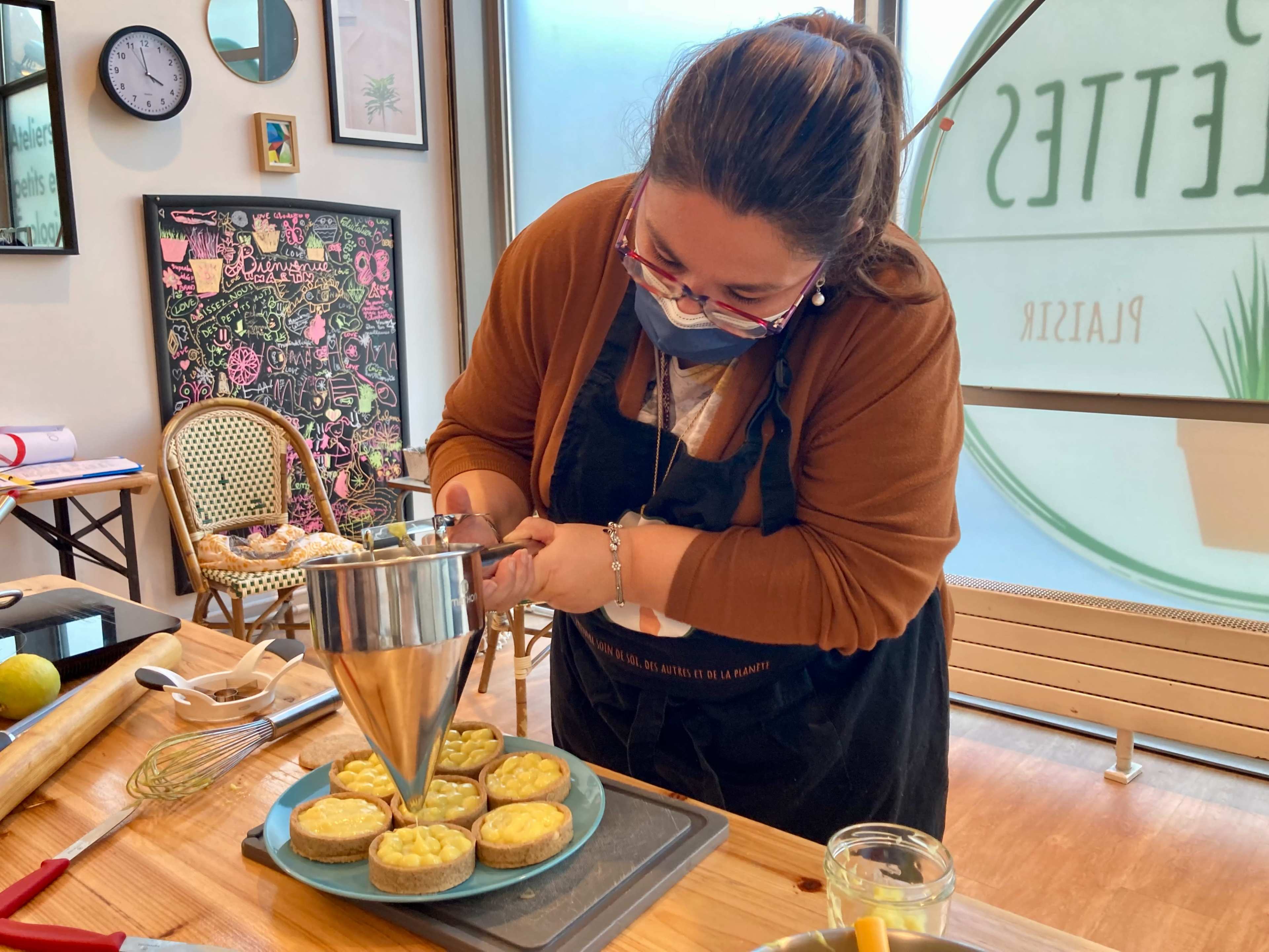 A woman wearing a mask is carefully piping filling onto small tarts in a kitchen setting.