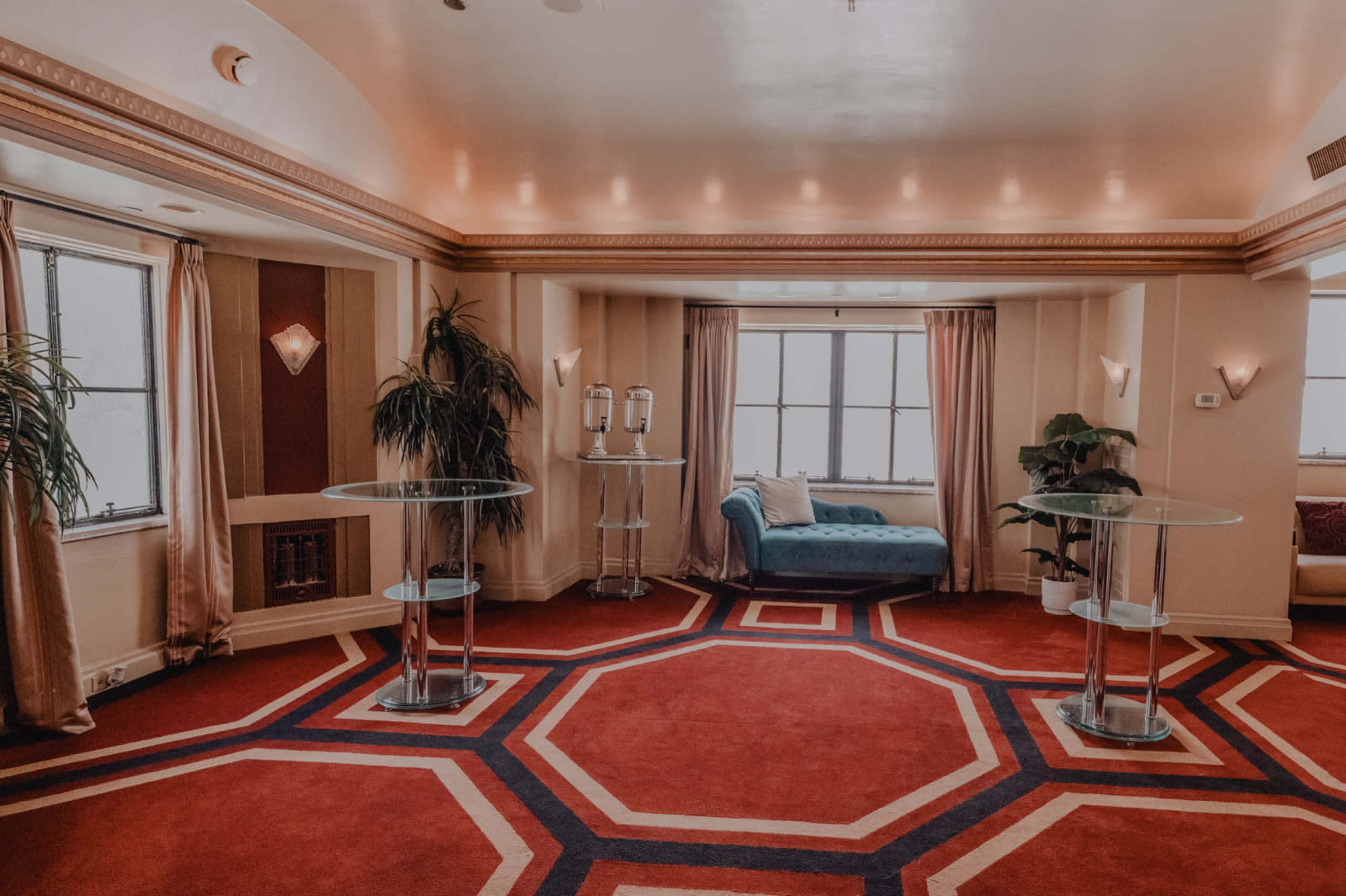 The image shows a spacious room with a red carpet featuring geometric patterns, clear glass tables, and a blue upholstered chaise lounge near large windows.