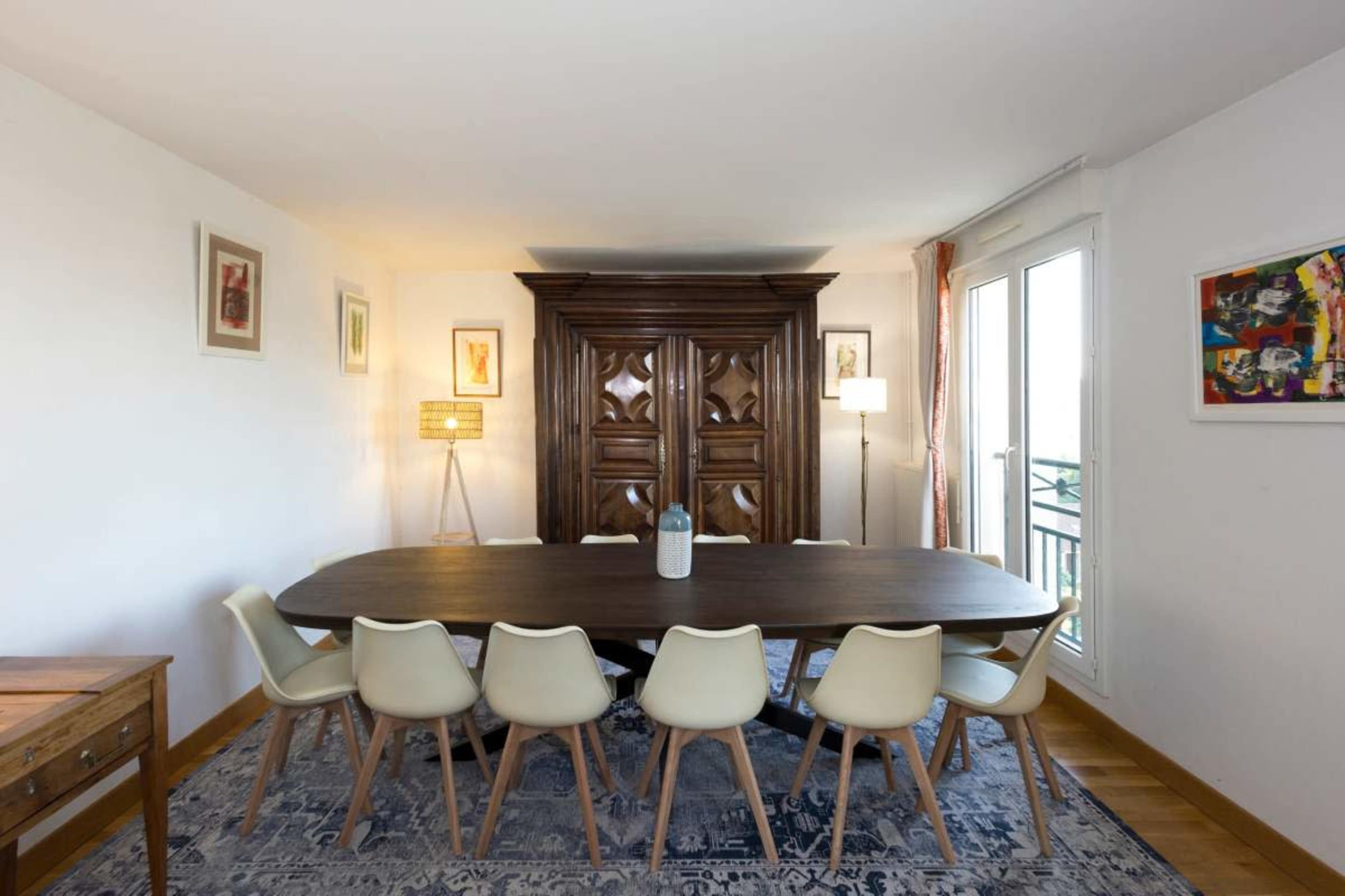 A spacious dining room features a long wooden table surrounded by white chairs, with a decorative cabinet and lamps providing ambient lighting.