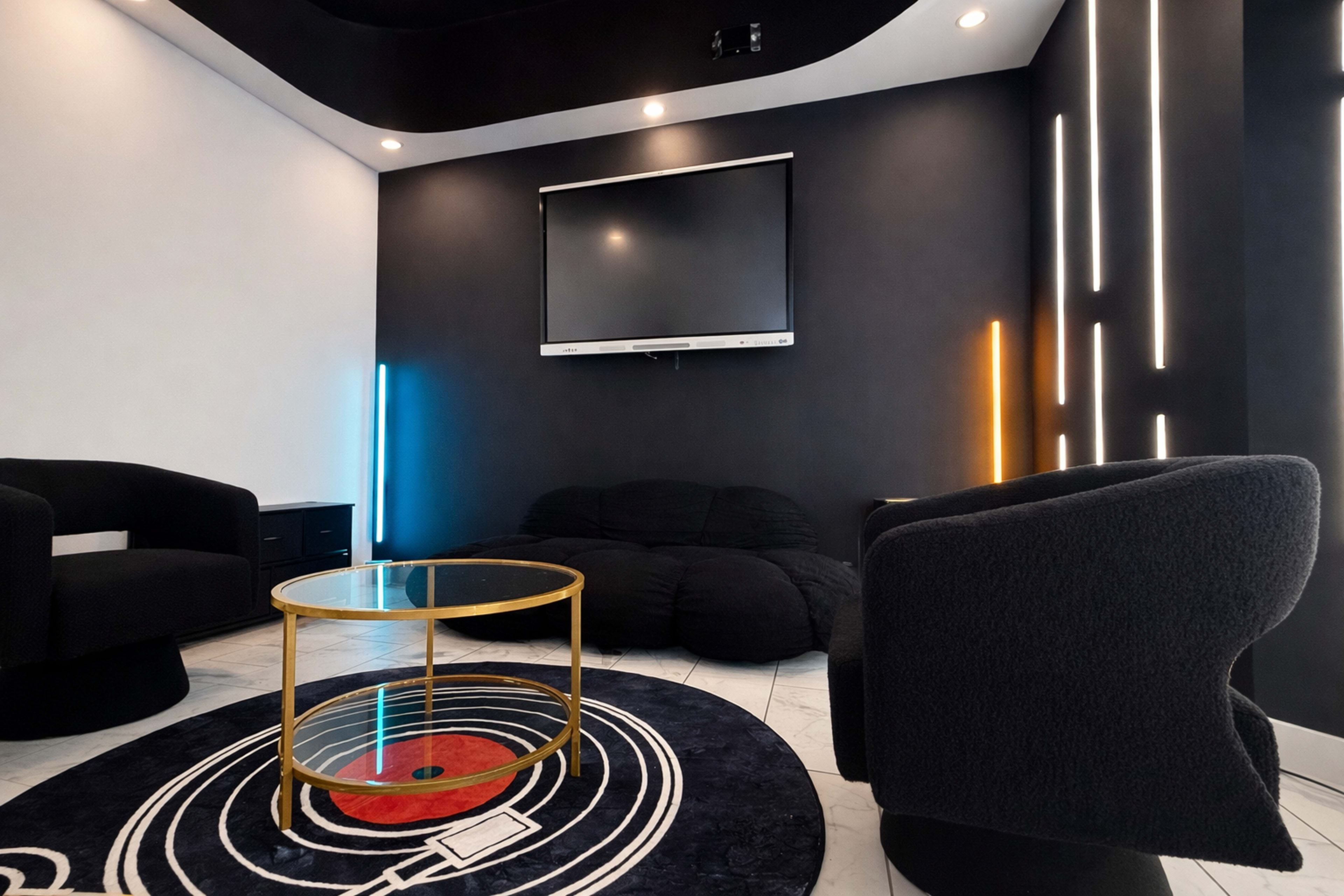 The image shows a modern lounge area featuring a low black seating arrangement, a round coffee table, and a wall-mounted television, with ambient lighting fixtures and a circular rug on the floor.