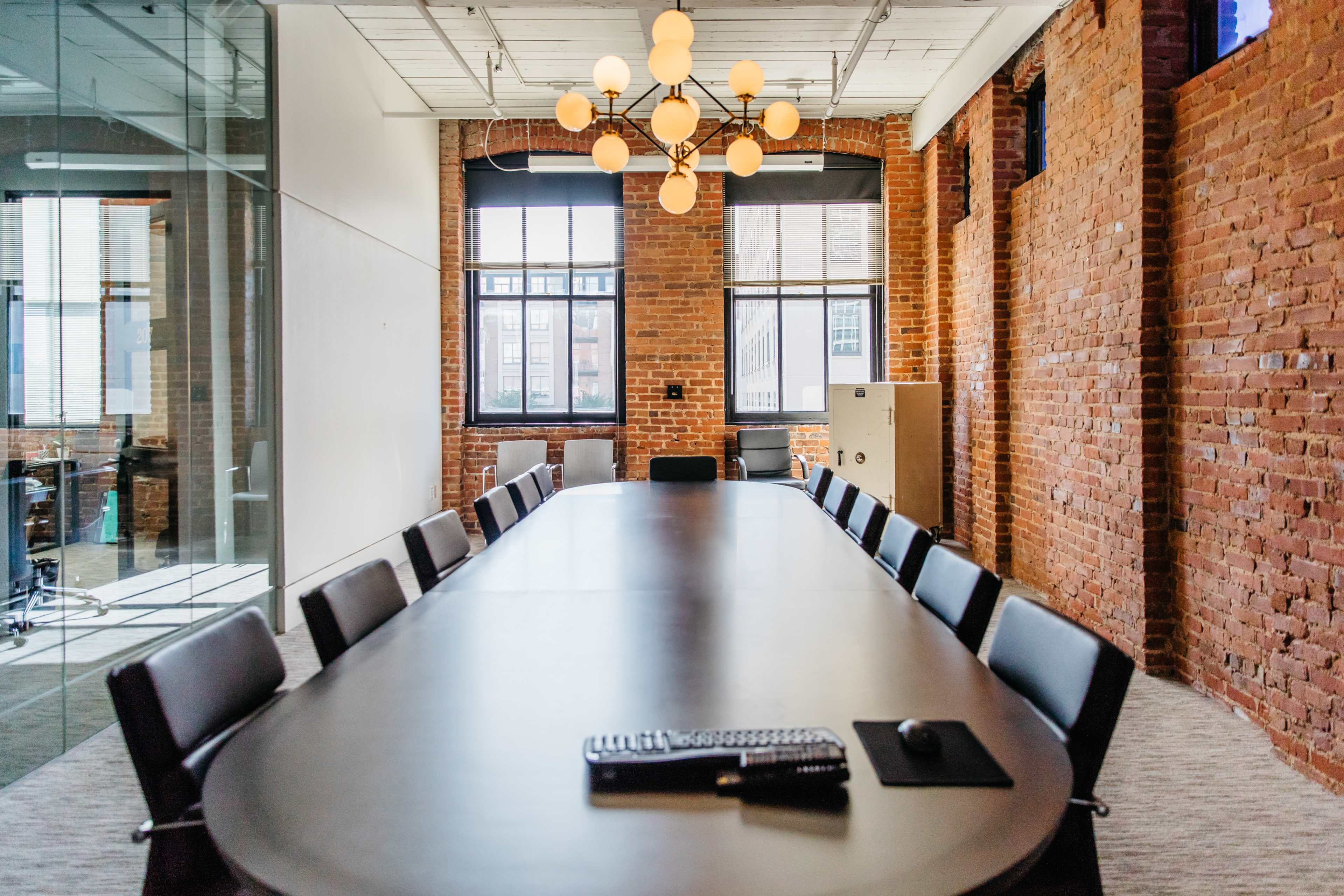 A long conference table with a computer keyboard and mouse sits in a room with exposed brick walls and large windows.