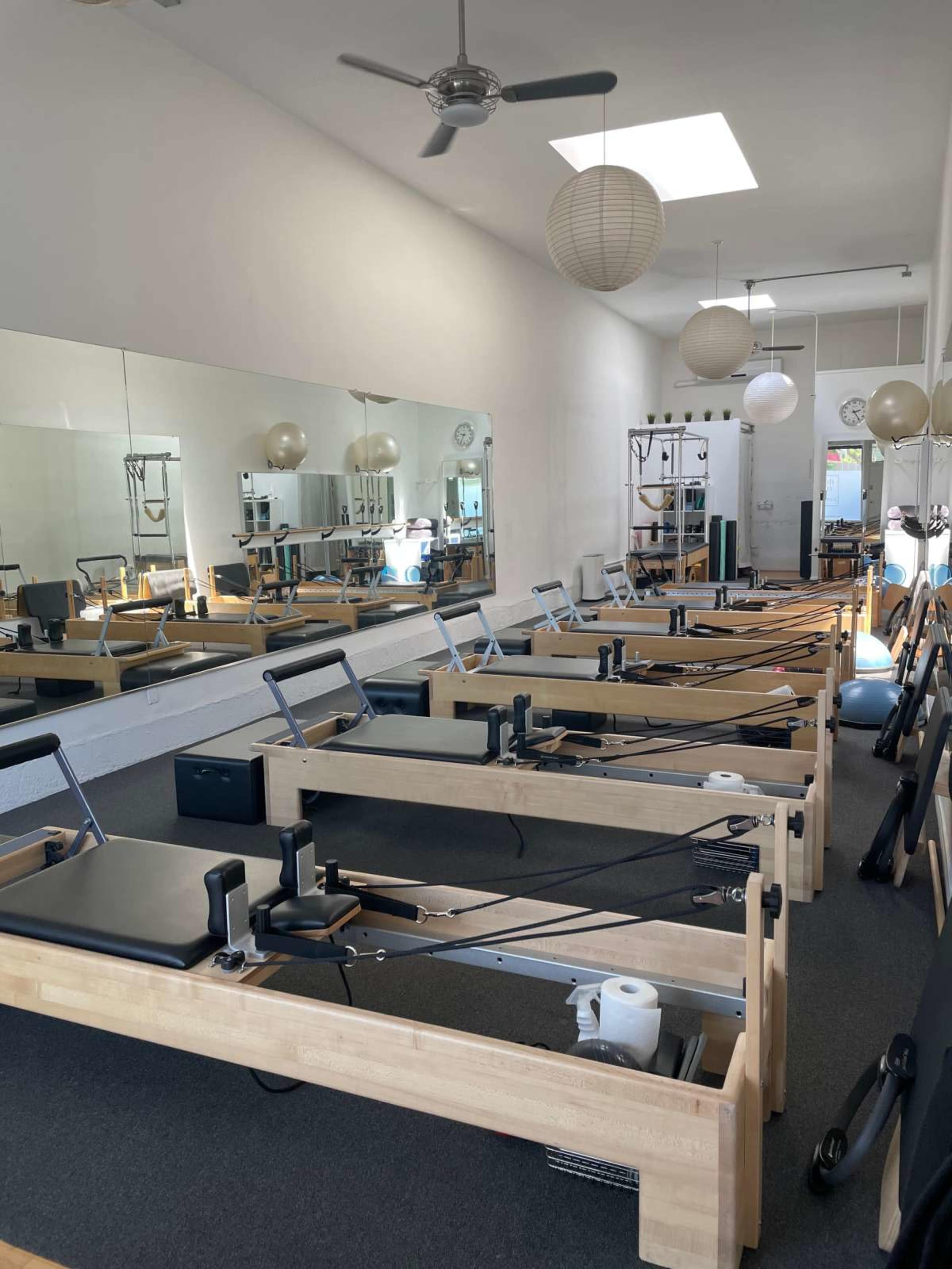 The image shows a bright Pilates studio filled with reformer machines, mirrors, and soft lighting.