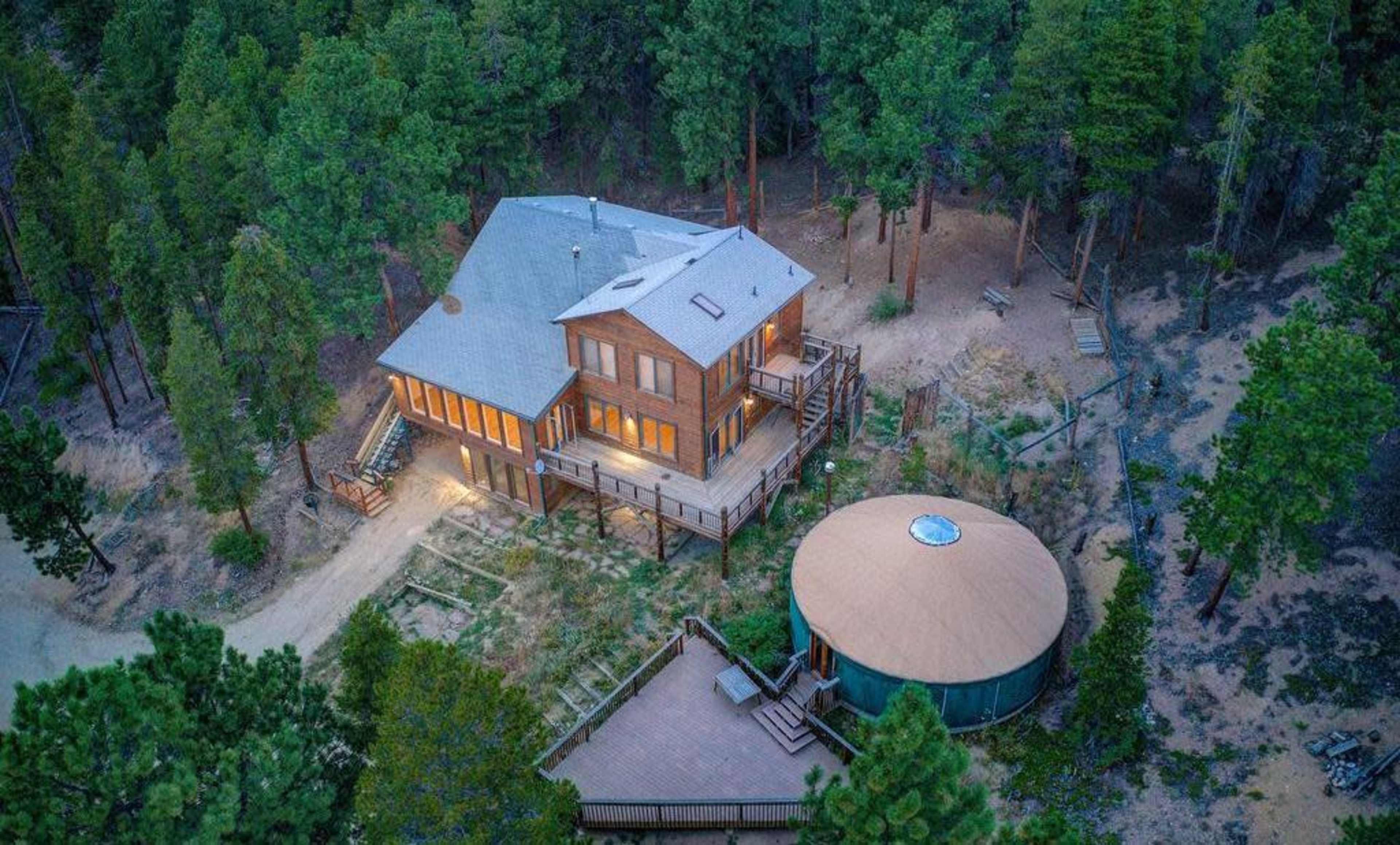 A large wooden house with multiple levels and large windows is surrounded by tall trees, with a circular structure and wooden deck nearby.