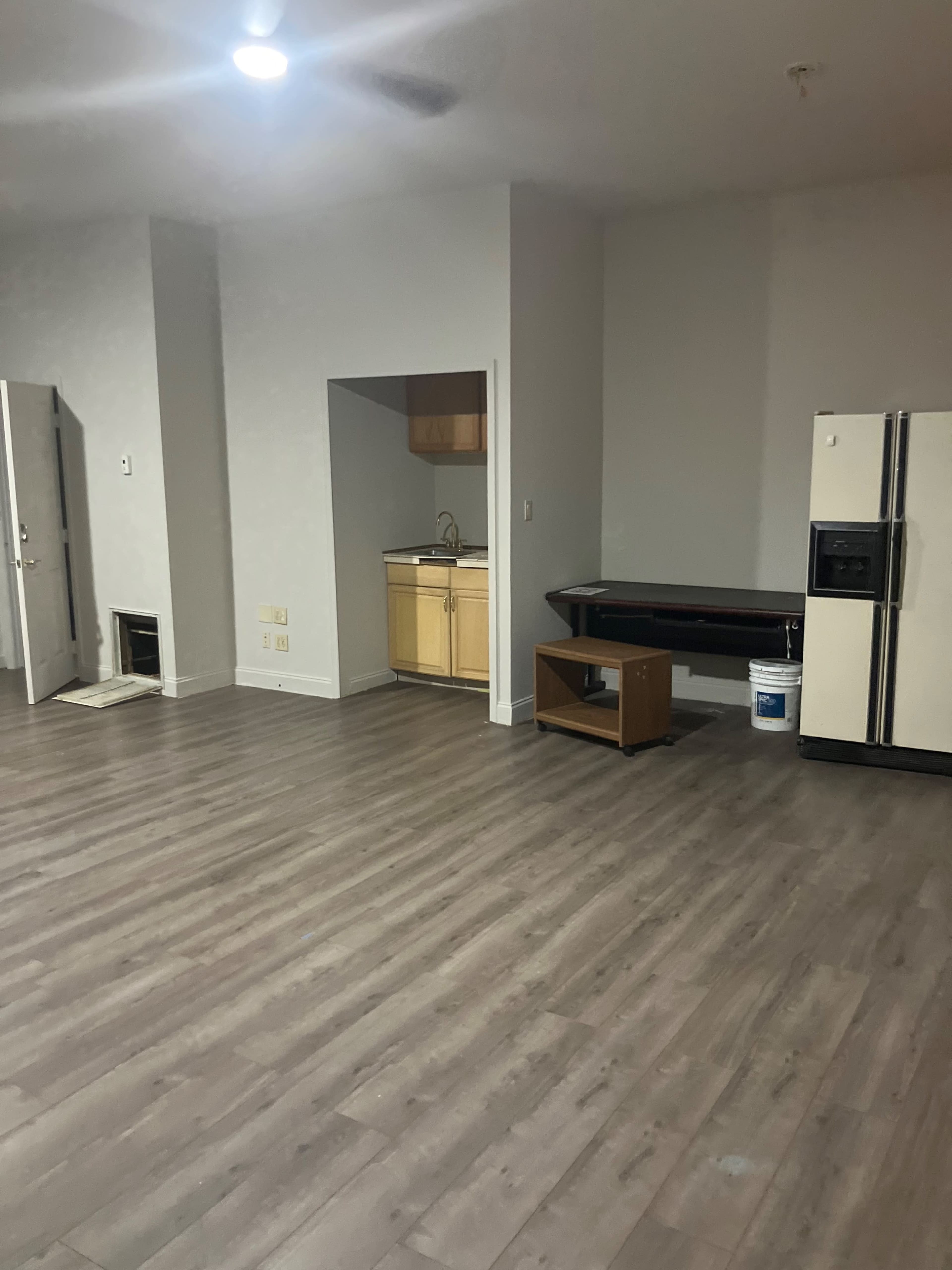 The image shows a spacious room with gray walls, a kitchen area in the back, a refrigerator on the right, and a table against the wall.