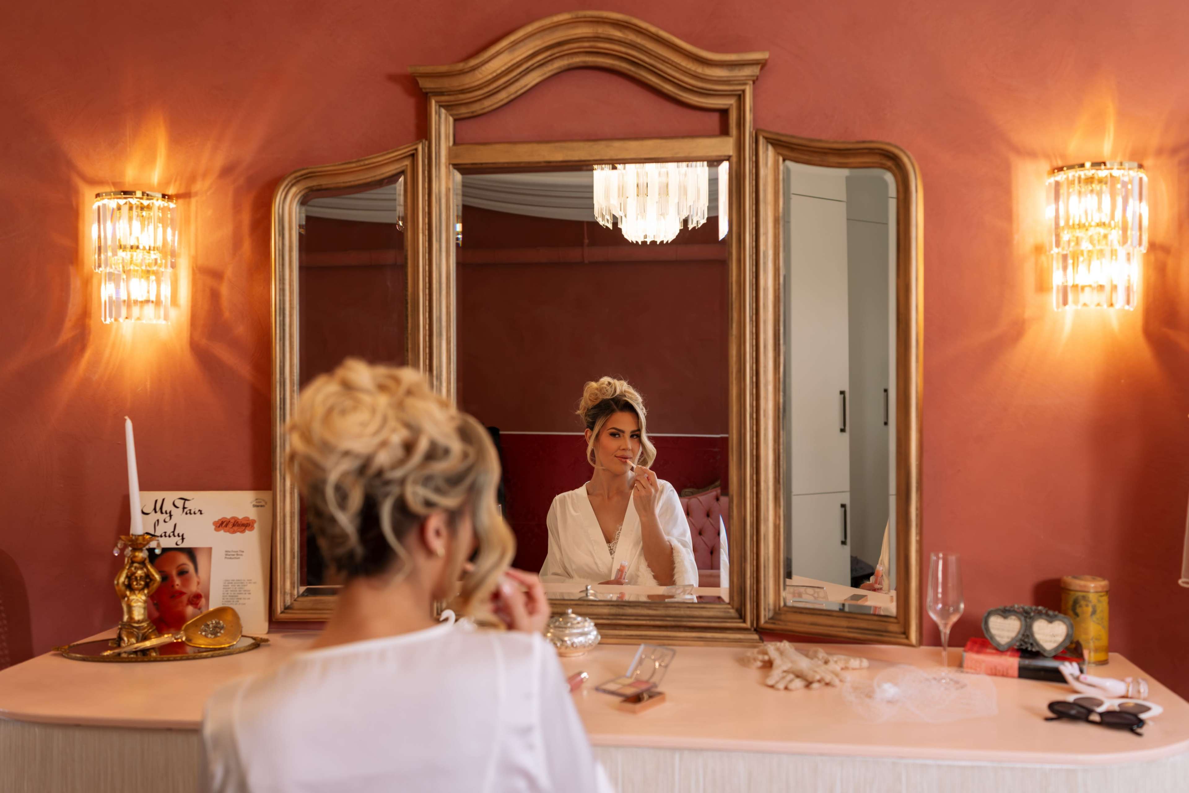 A woman sits at a vanity with elegant mirrors, applying makeup in a softly lit room adorned with warm-colored walls and decorative accents.
